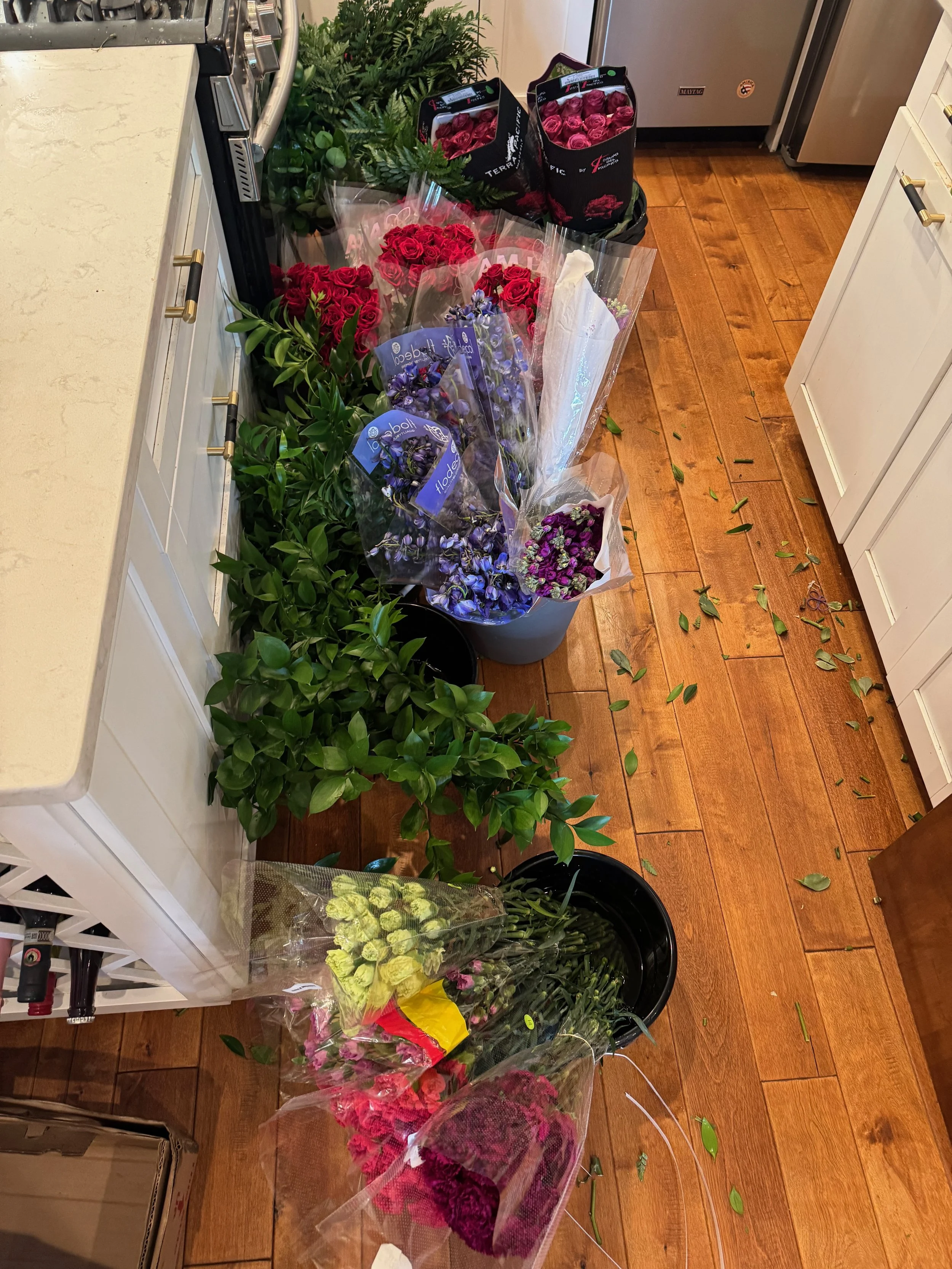 Various flower bouquets and potted plants on a hardwood kitchen floor, with some leaves scattered around.
