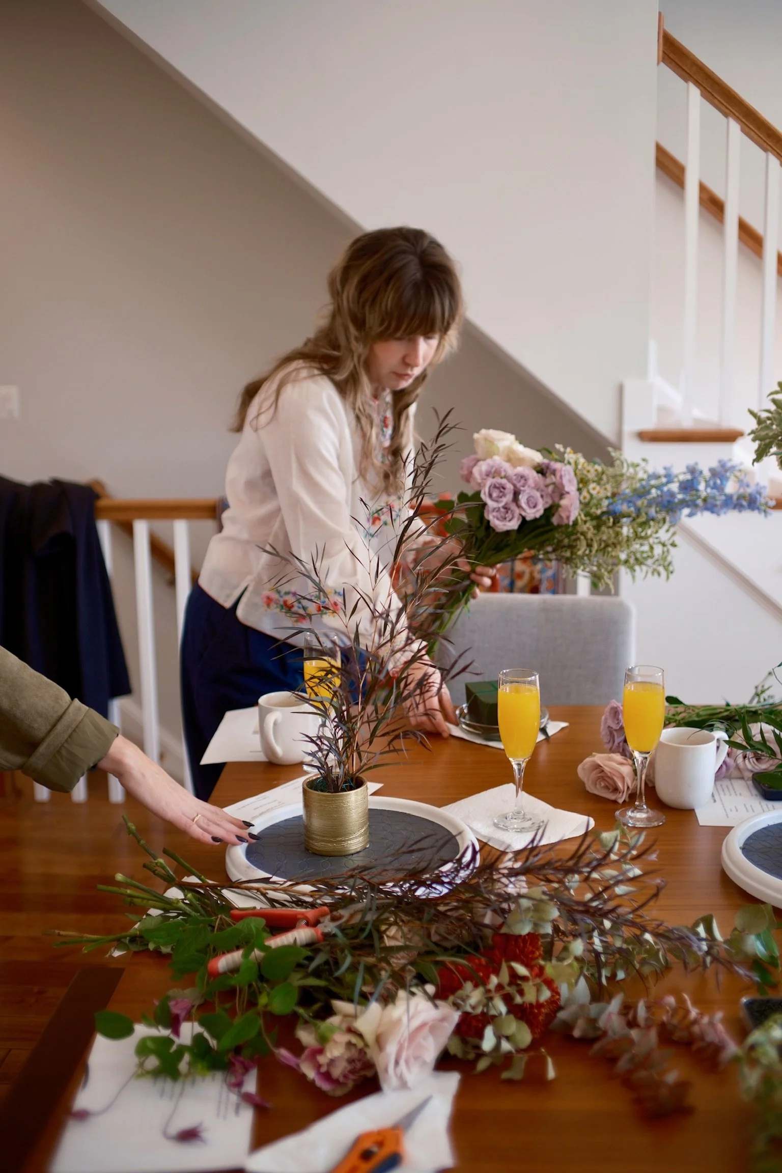 A woman arranging flowers on a dining table with floral arrangements, glasses of orange juice, and a notebook.