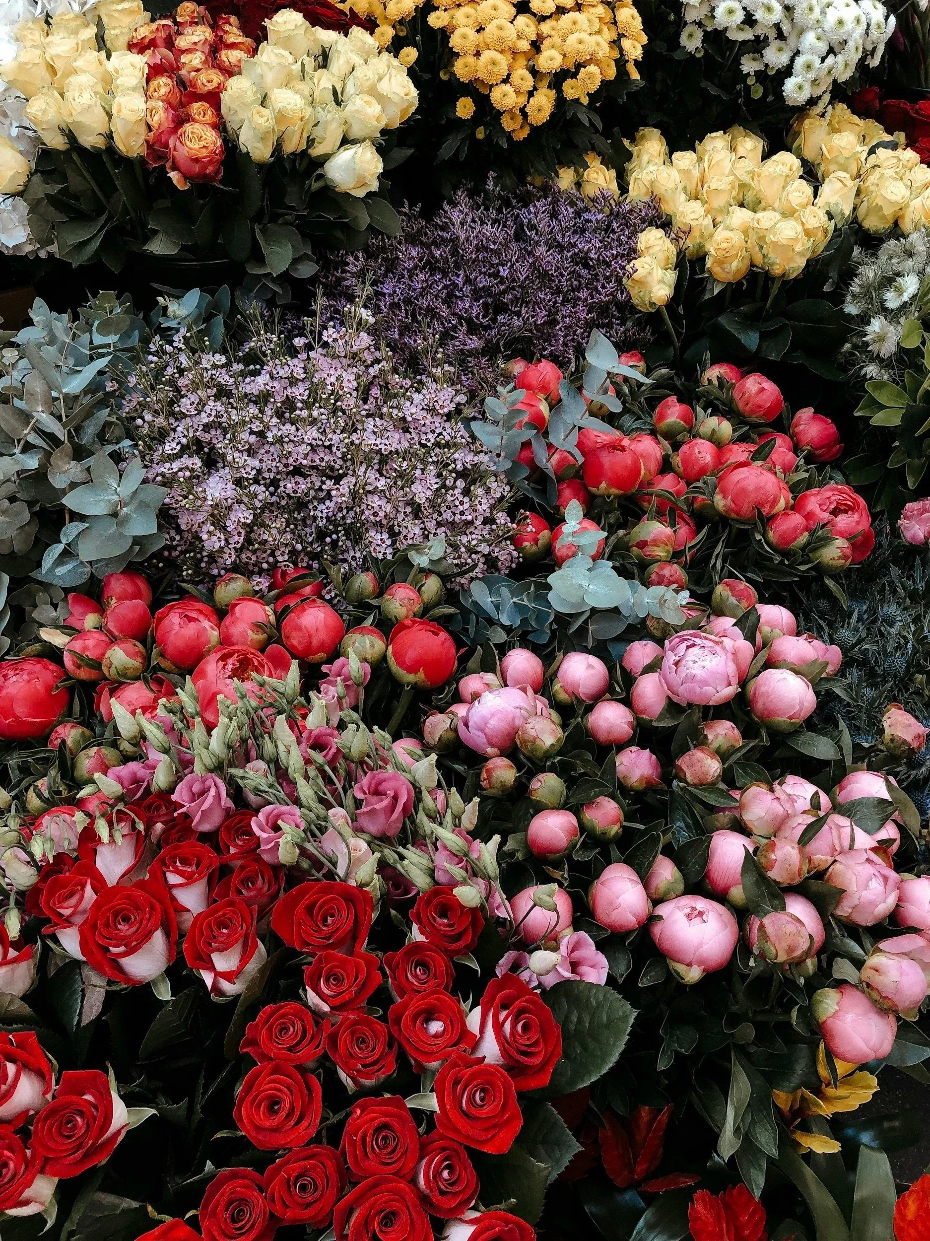 A colorful display of various flowers including roses, peonies, and other blooms in shades of red, pink, purple, white, yellow, and lavender, arranged closely together.