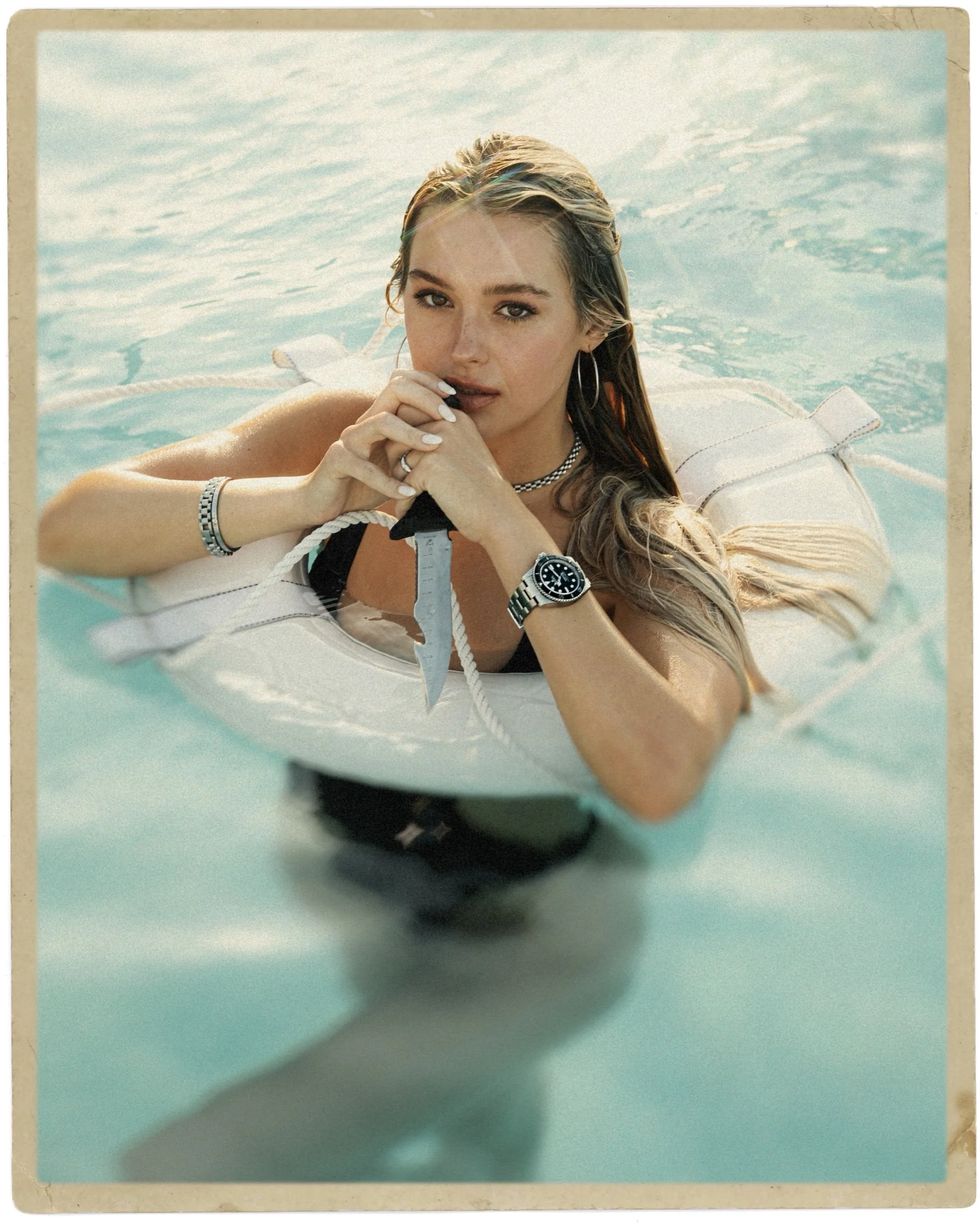 Young woman with wet hair lying on a boat or watercraft, holding onto a rope, with a chain necklace, hoop earrings, and a wristwatch, in a body of water on a sunny day.