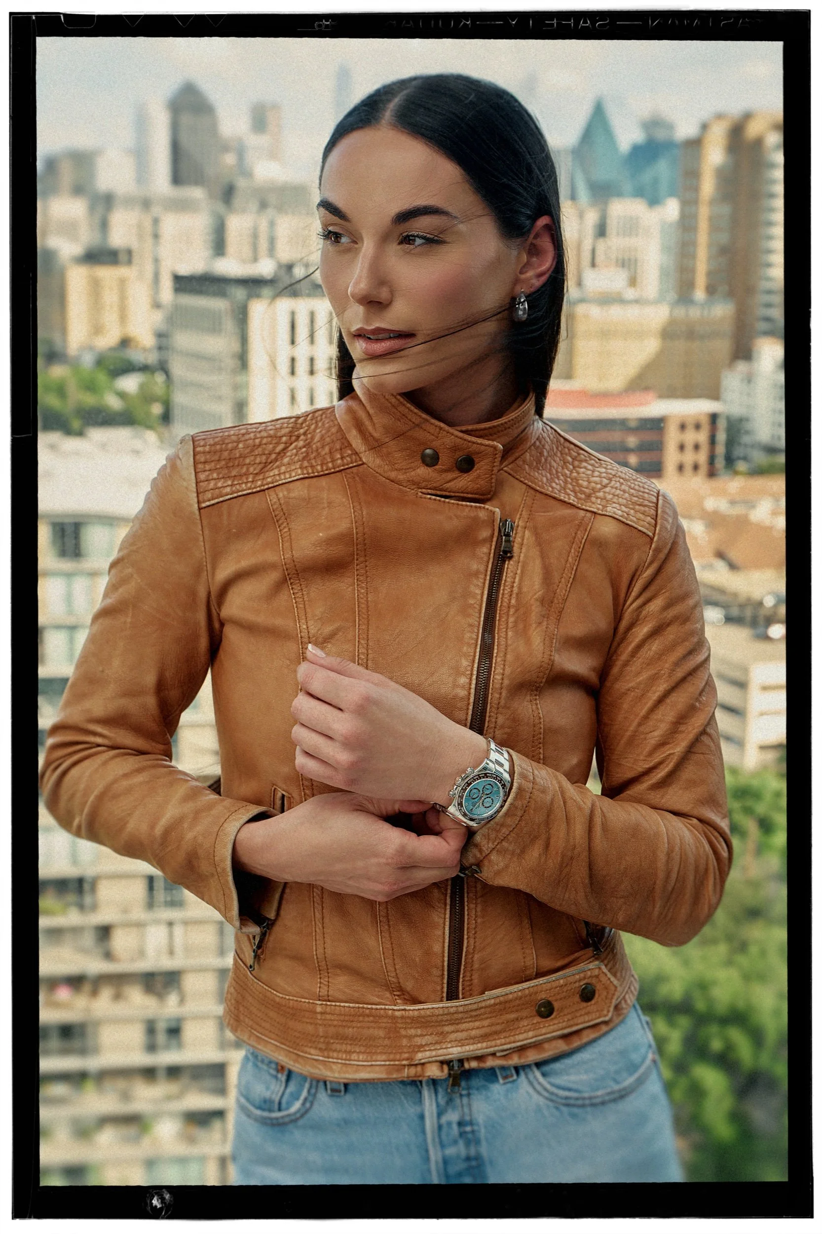 A woman with dark hair wearing a brown leather jacket and blue jeans standing outdoors in front of a cityscape with tall buildings.