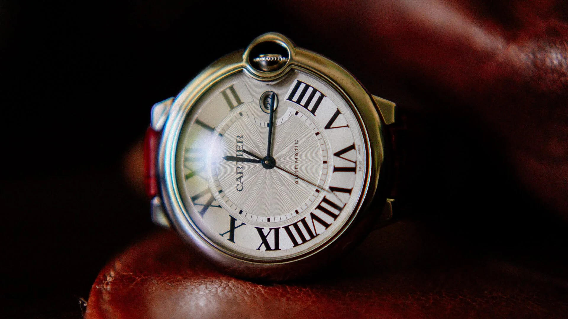 Close-up of a Cartier ballwatch with Roman numerals, showing 12:00, on person's wrist with brown leather strap.