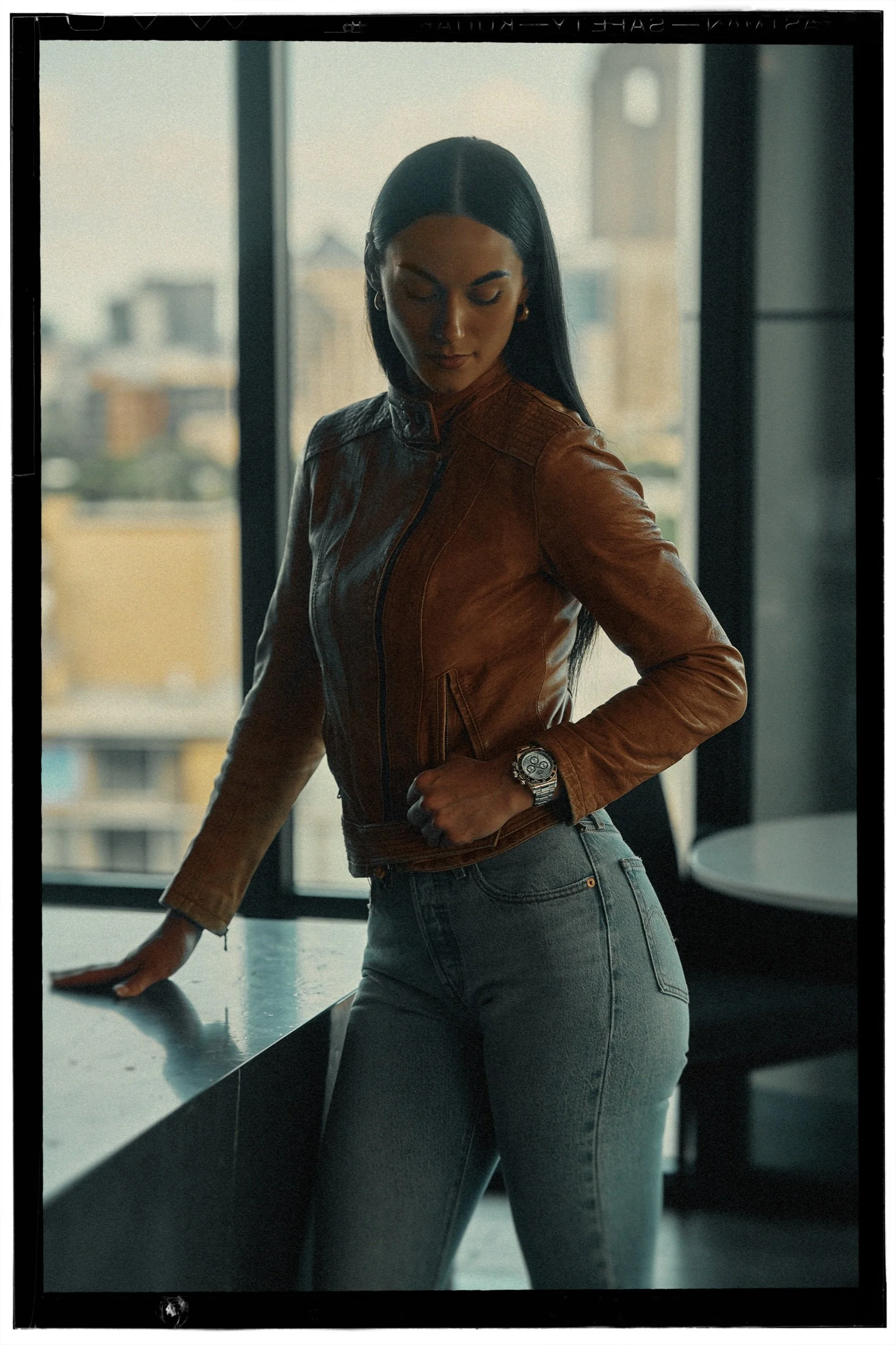 A woman with long dark hair is standing by a black countertop, wearing a brown leather jacket and gray jeans. She is looking down with a slight smile, in a modern room with large windows showing city buildings in the background.