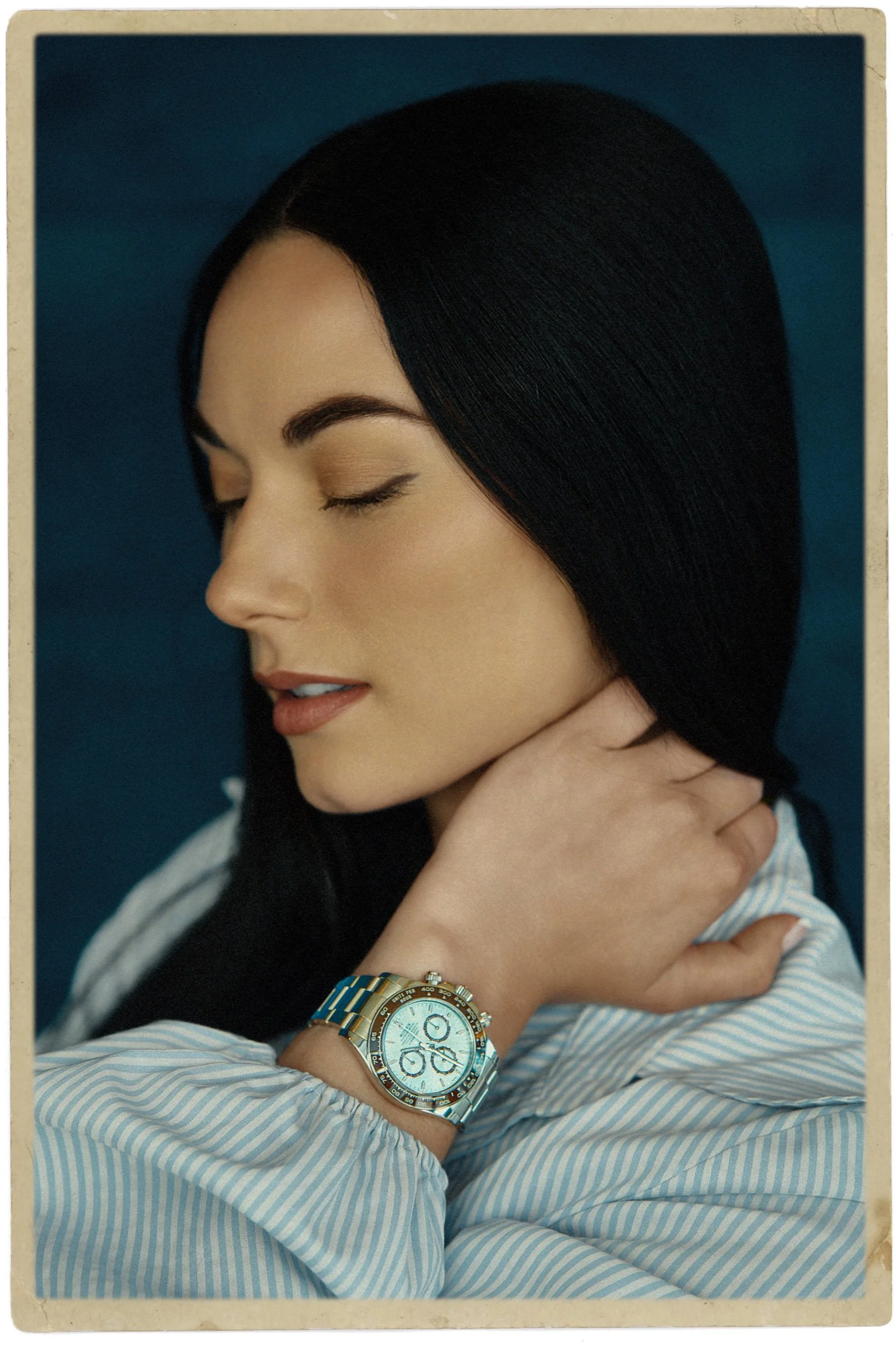 A woman with black, straight hair and makeup, wearing a striped shirt and a large gold watch, is resting her chin on her hand with her eyes closed.