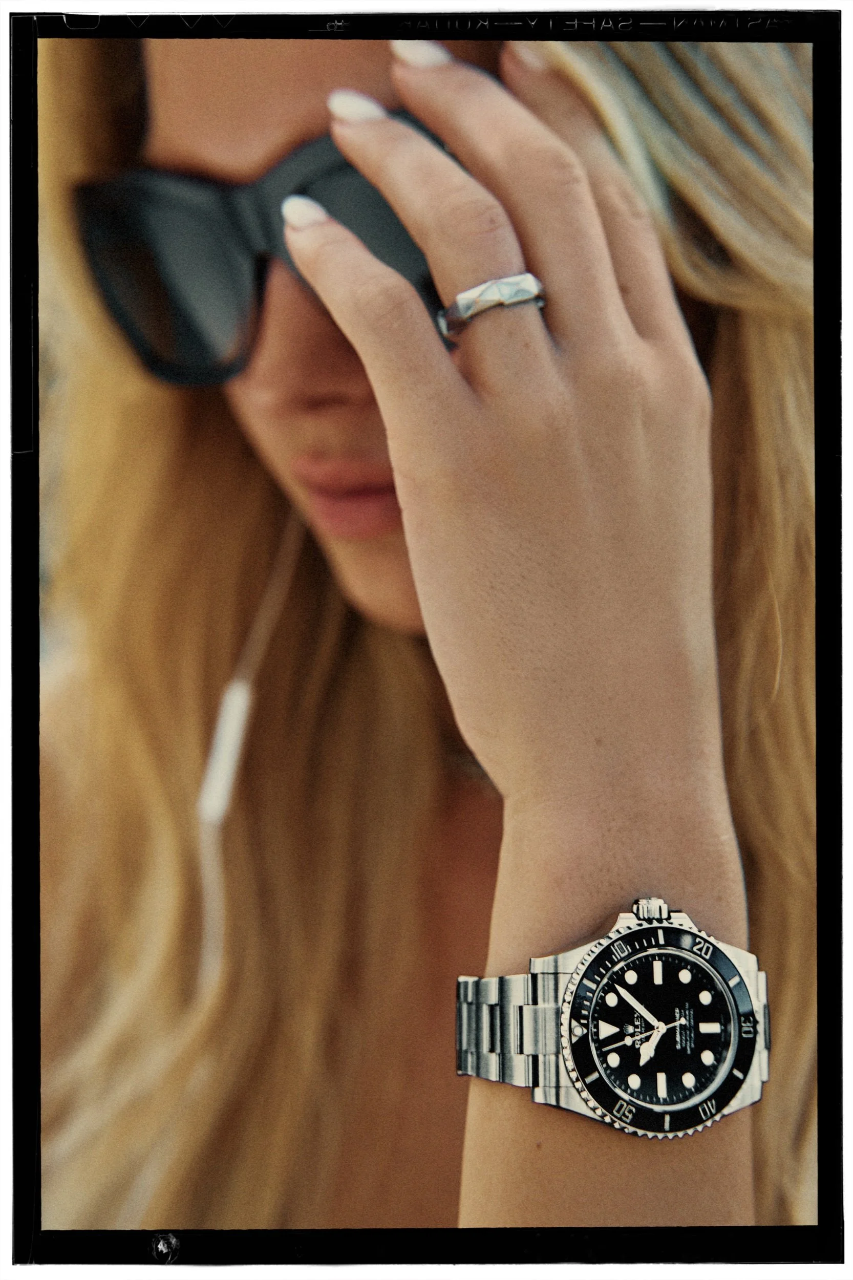 Close-up of a woman with long blonde hair wearing a silver wristwatch and a silver ring, holding her hand near her face with a black watch or eyewear.