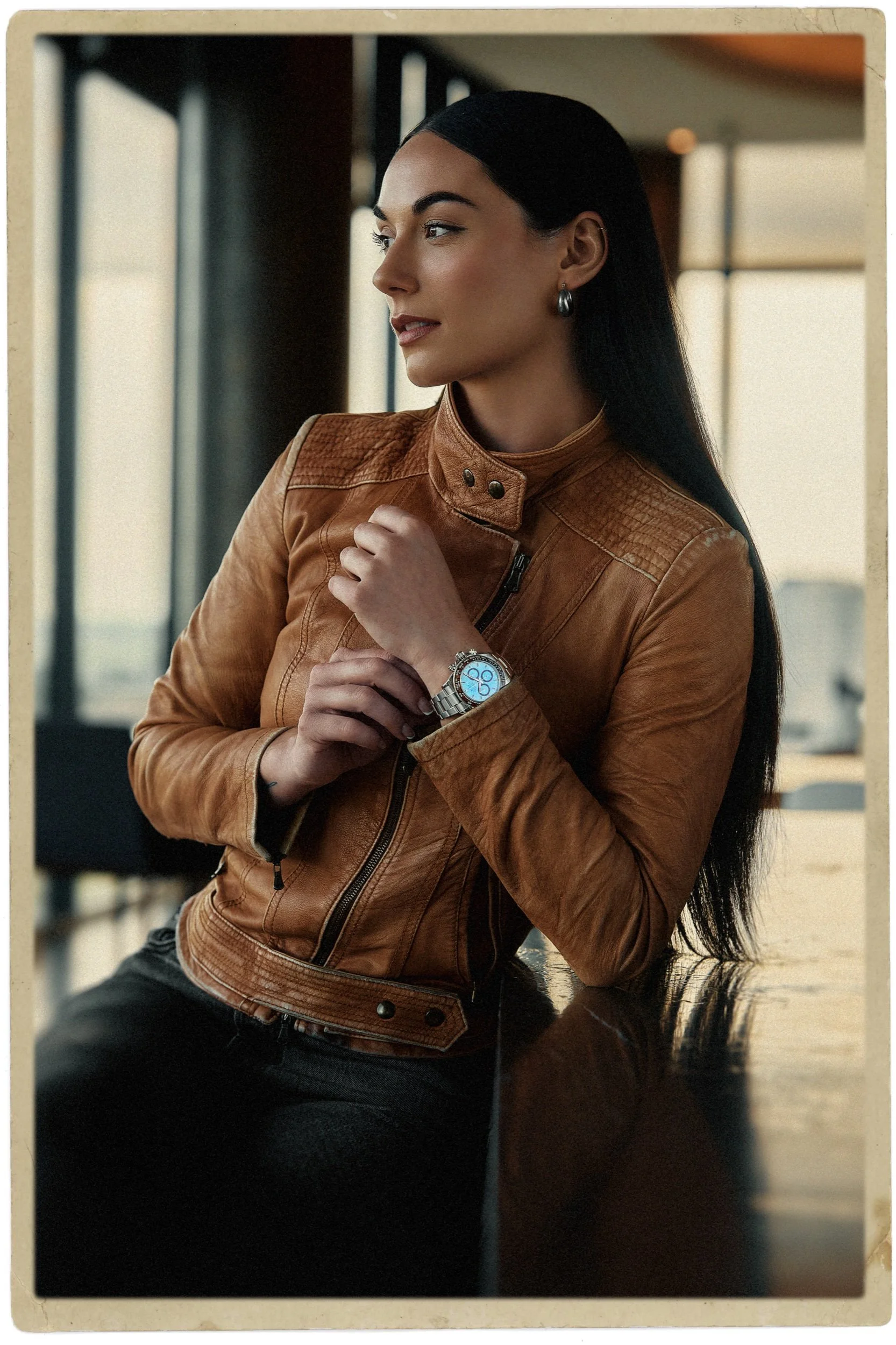 A woman with long black hair wearing a brown leather jacket, earrings, a watch, and black pants, sitting indoors near large windows.