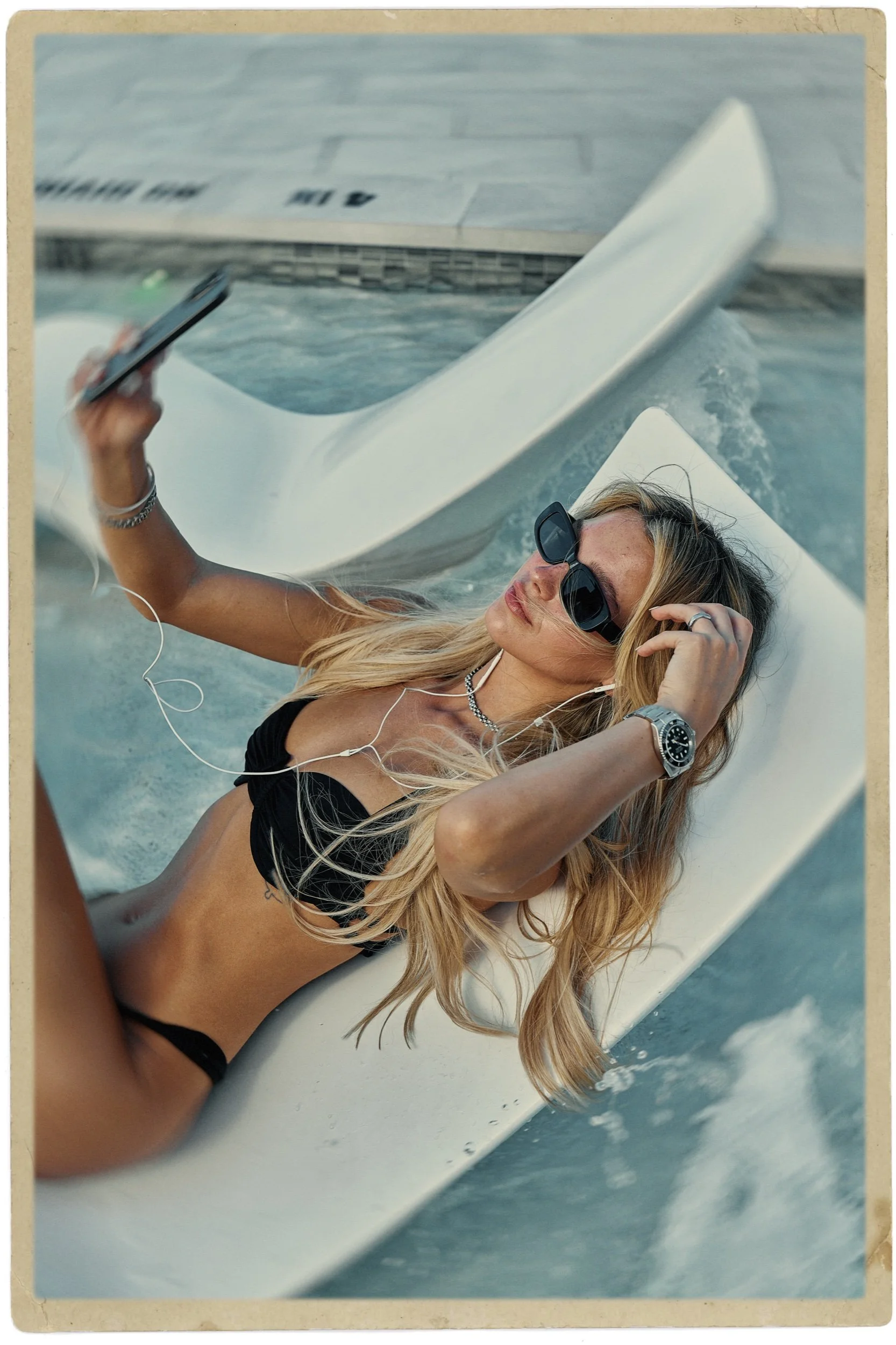 A woman in black bikini lying on a surfboard, holding her phone, with sunglasses and headphones, on a boat or water vessel.
