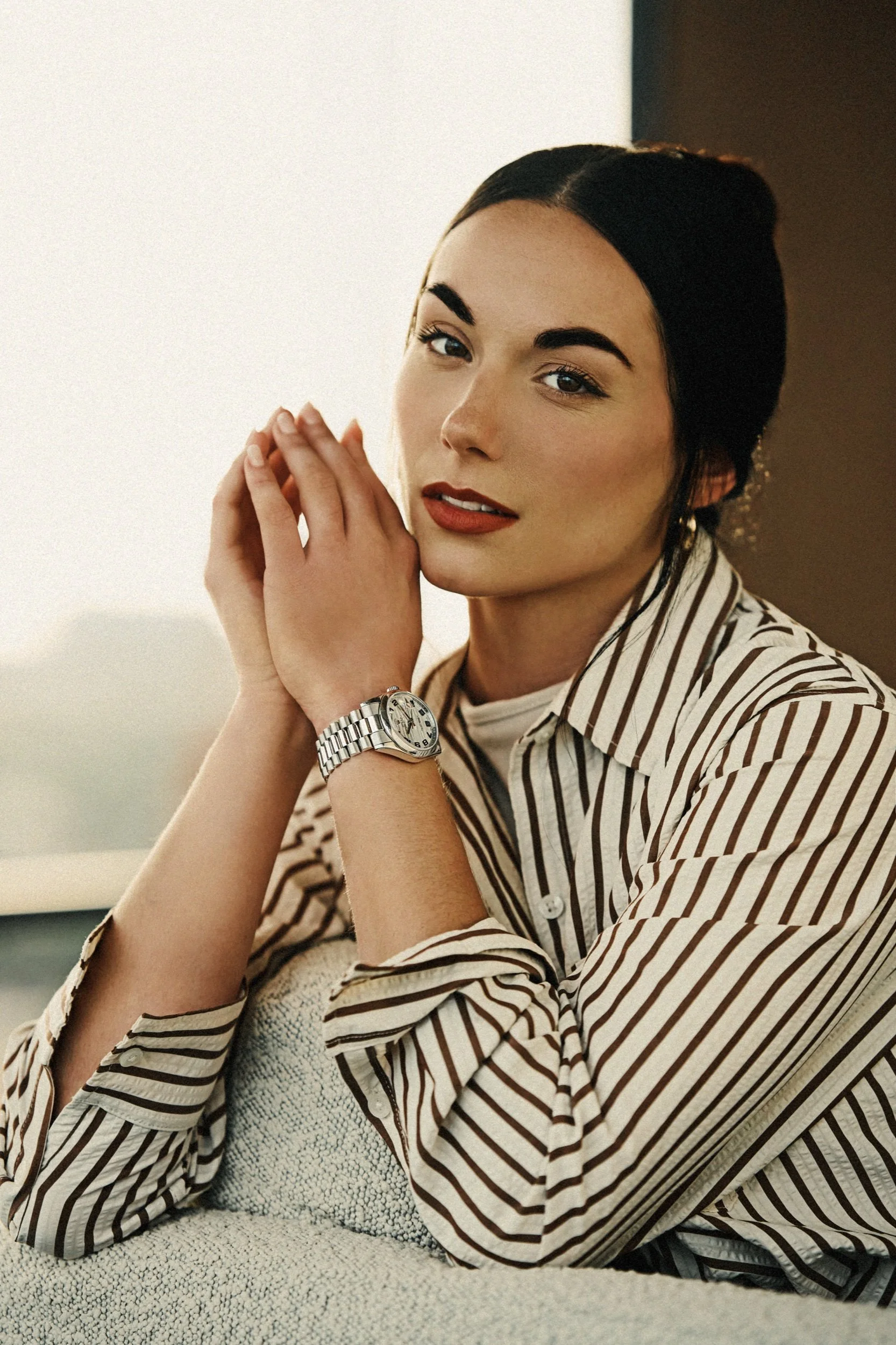 A woman with dark hair and makeup, wearing a striped shirt and a watch, rests her chin on her hands while looking at the camera, sitting on a cushioned surface in front of a plain background.