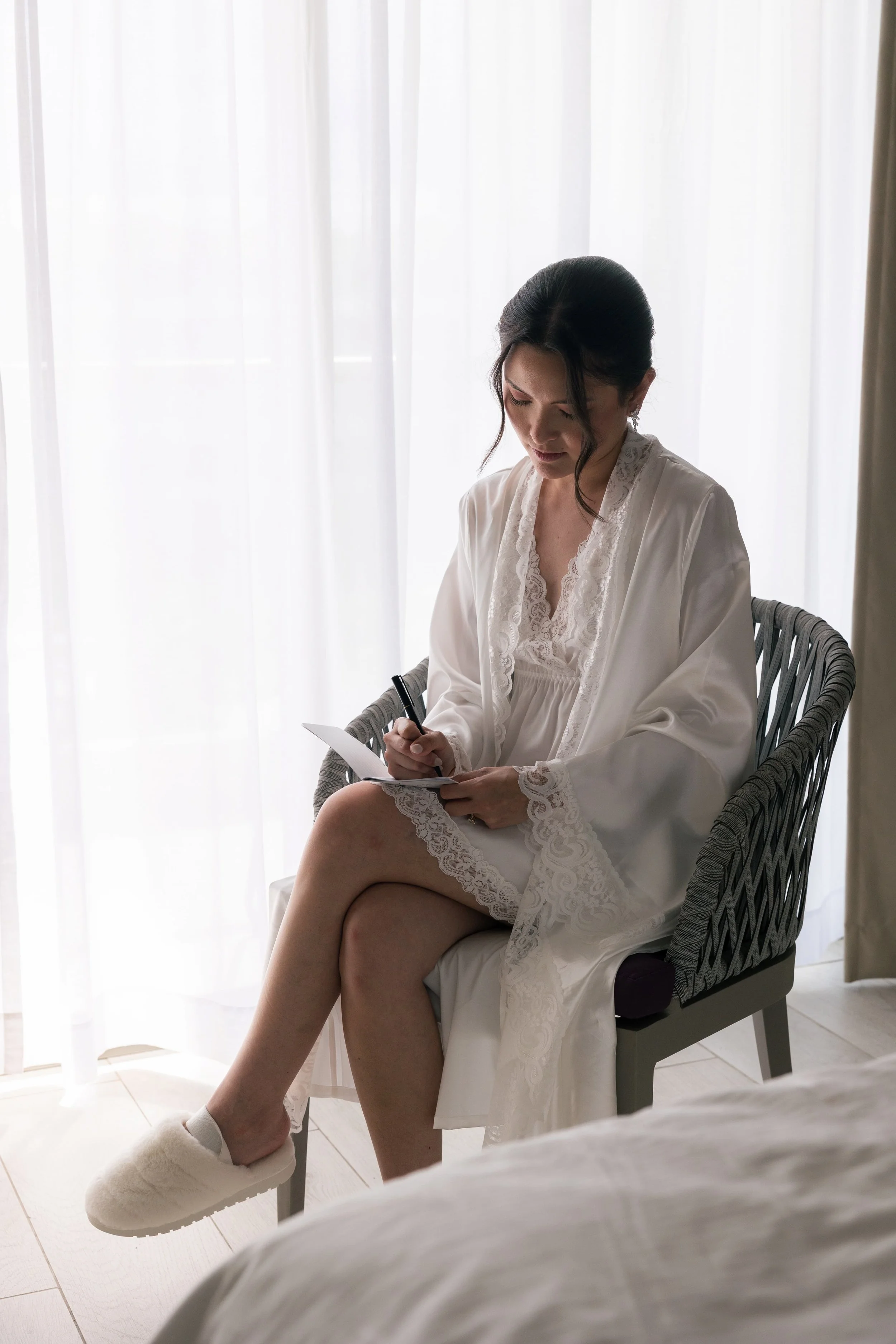 A woman in a white robe sitting on a chair near a window with sheer curtains, writing on a notepad.
