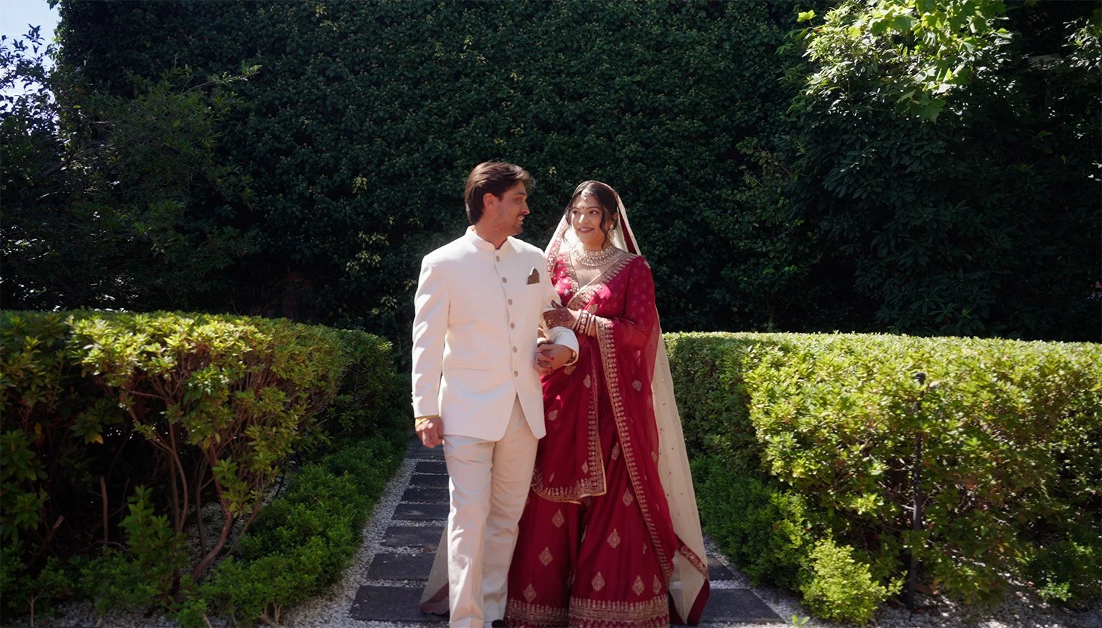 A couple dressed in traditional Indian wedding attire walking outdoors amid greenery, with the woman smiling at the man.