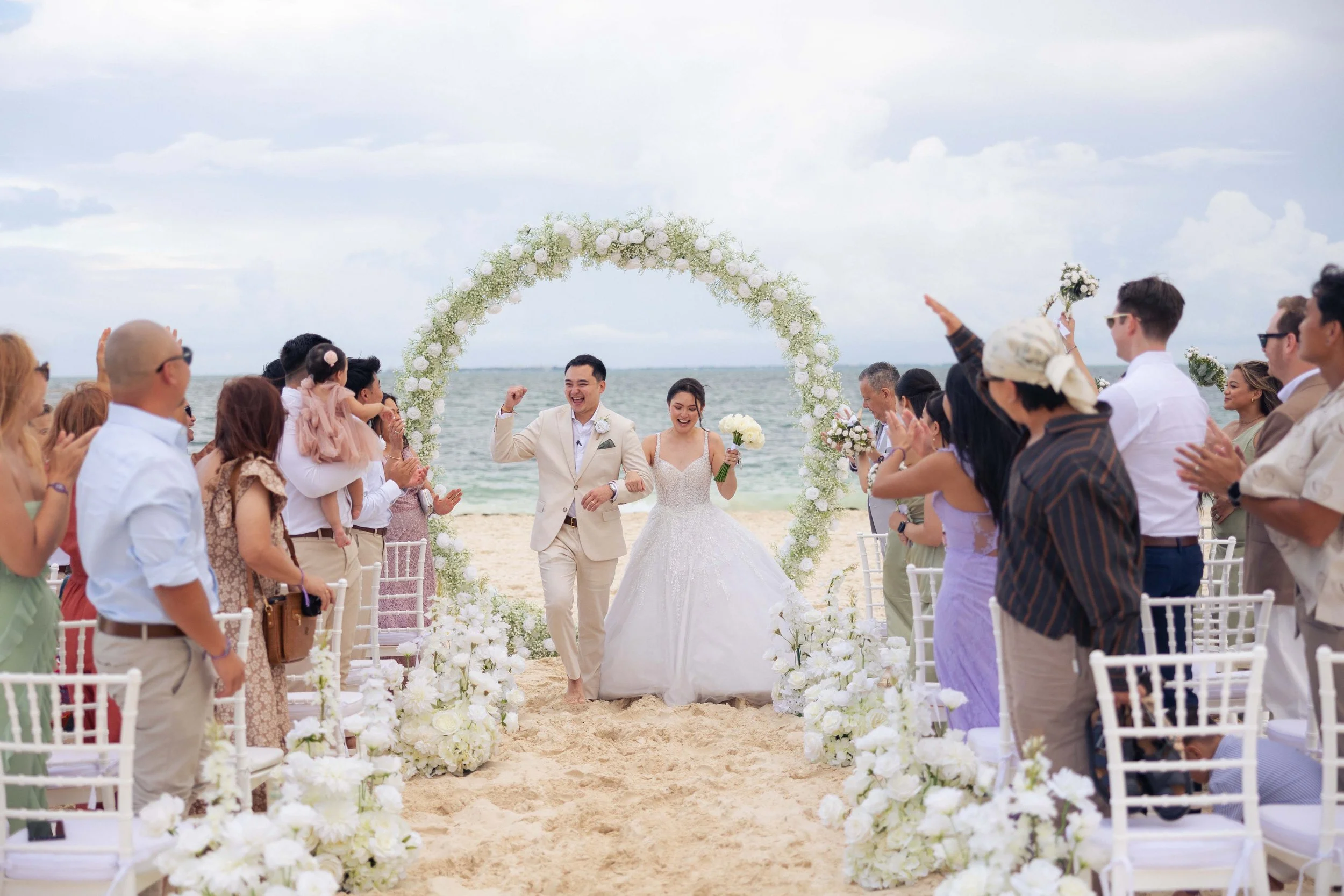 A newlywed couple walks down the aisle on a beach, smiling and celebrating after their wedding ceremony, surrounded by friends and family, with a floral arch in the background.