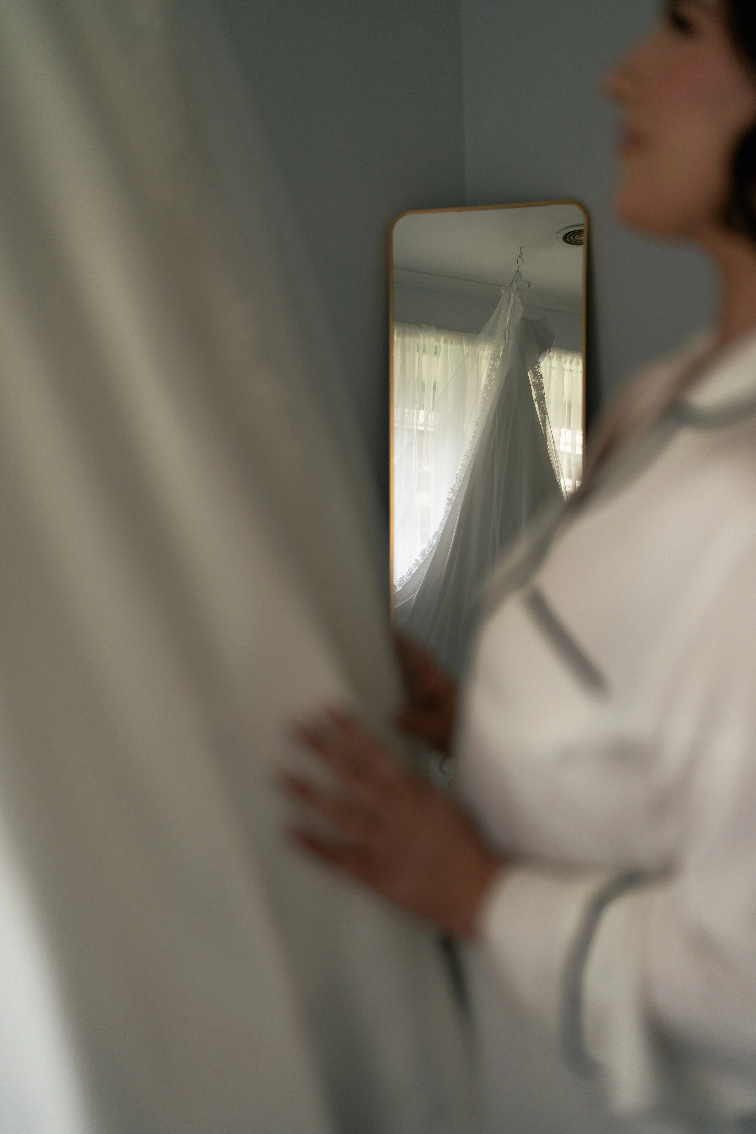 A woman looking into a mirror, with the reflection showing a wedding dress hanging on a window curtain.