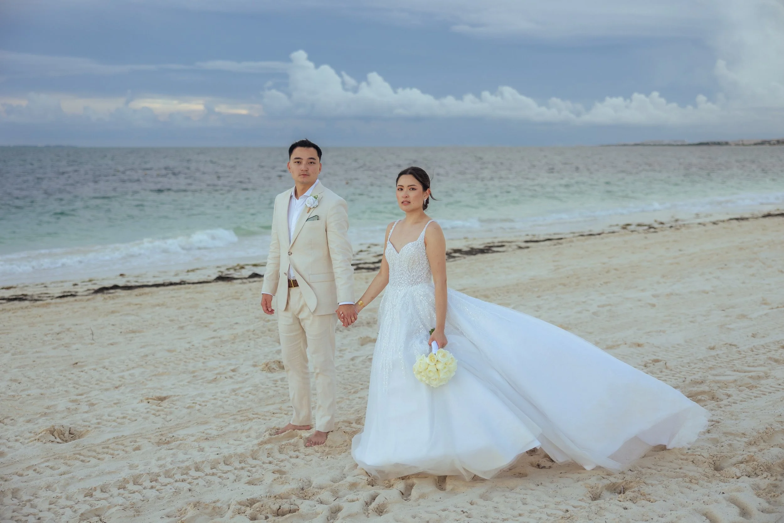 Bride and groom in wedding attire holding hands on a beach with ocean and cloudy sky in the background.