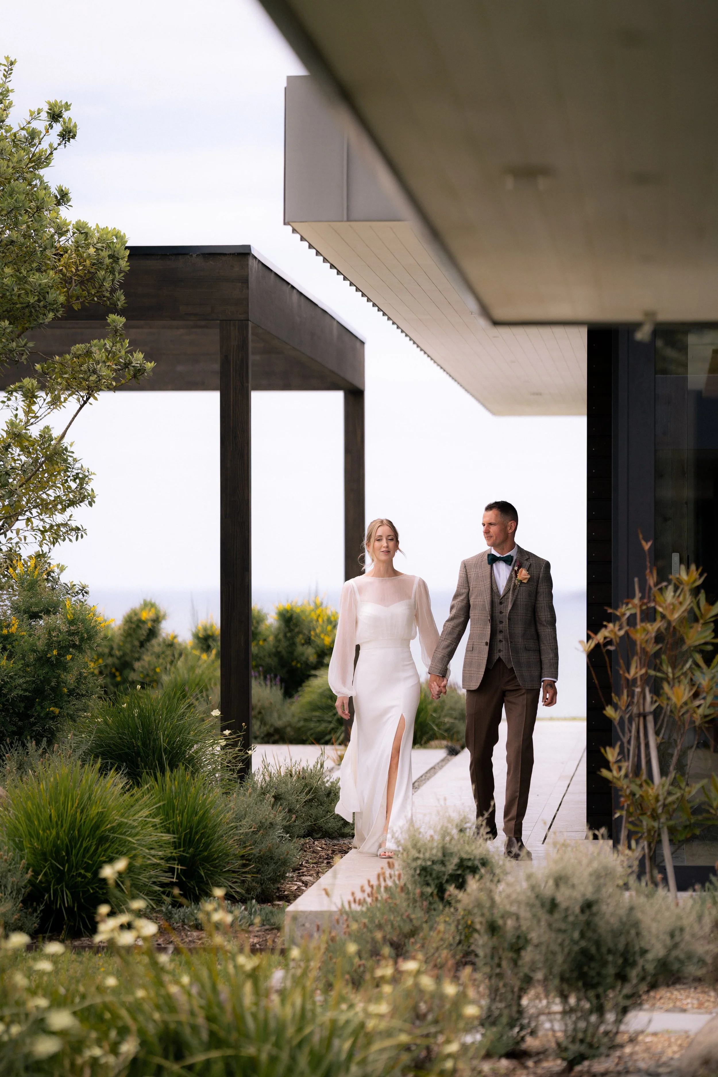 A bride and groom holding hands and walking outdoors near a modern building with lush greenery around them.