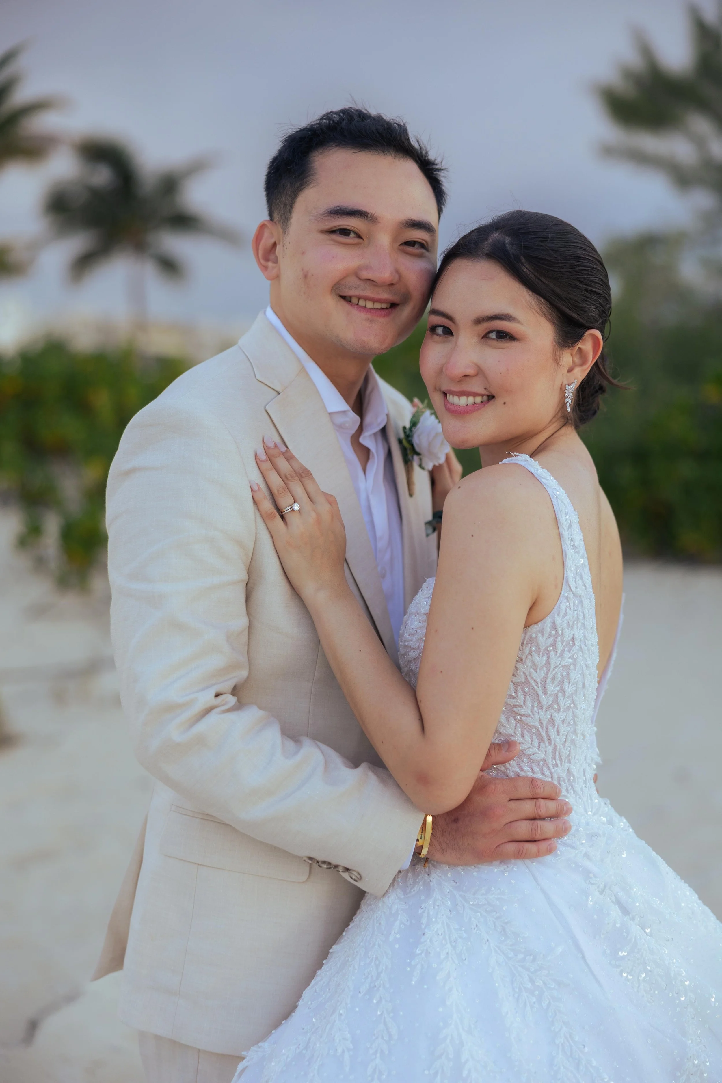 A newlywed couple embraces on a beach, with palm trees in the background. The groom wears a cream suit, and the bride wears a white lace wedding gown. They smile happily at the camera.