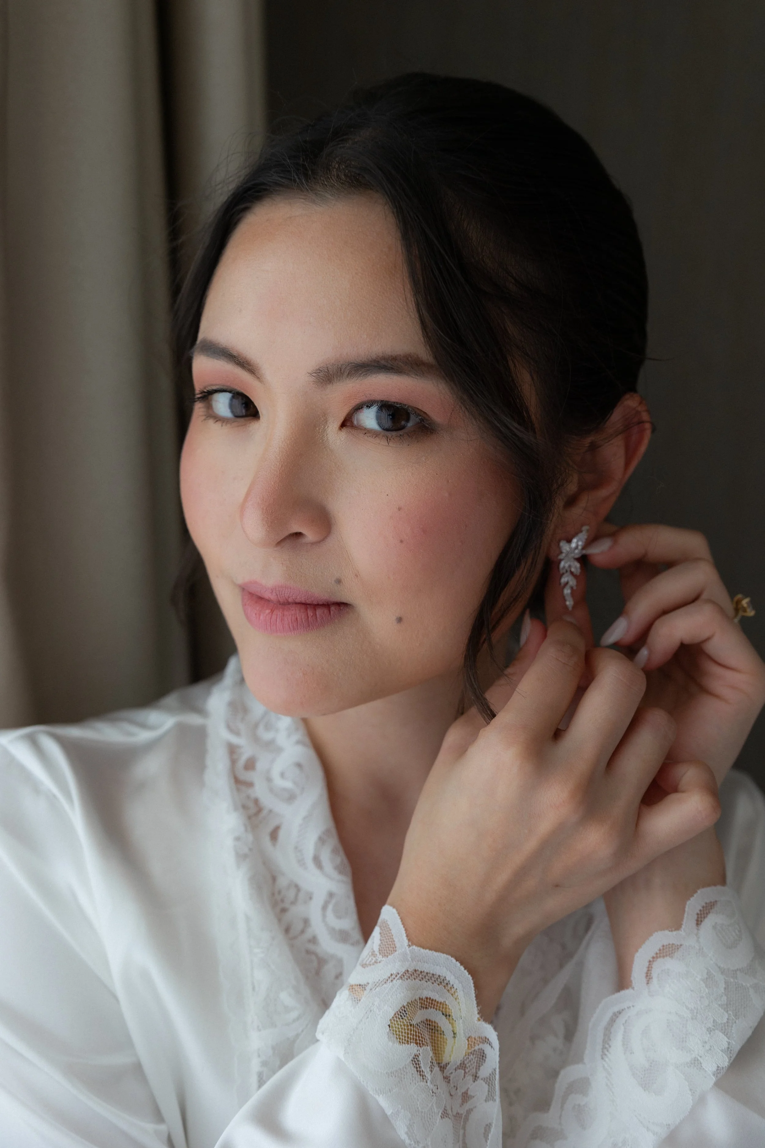A woman with dark hair styled back, wearing a white lace robe, adjusting an ornate diamond earring.