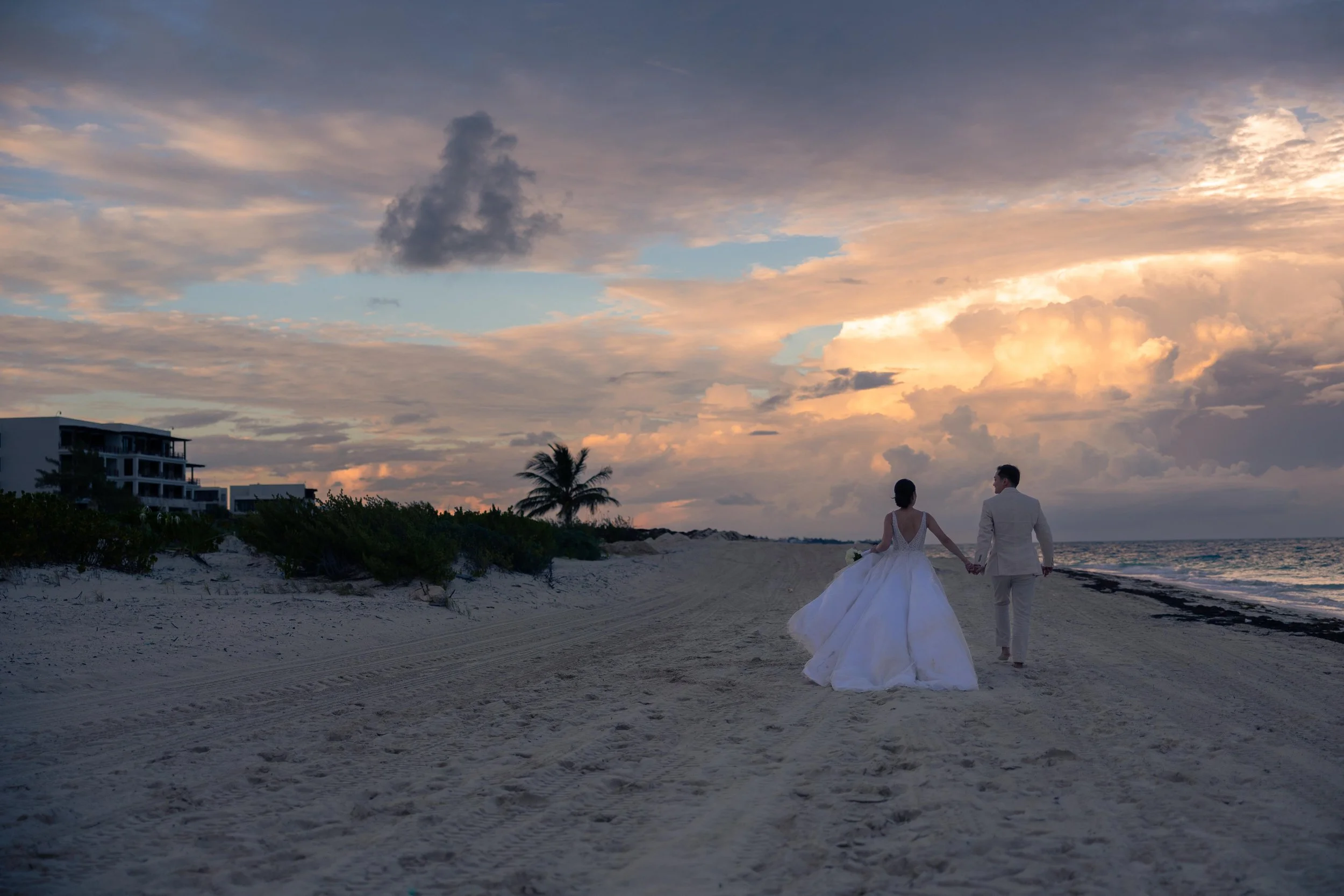 A bride and groom walking hand in hand on a sandy beach at sunset, with clouds and a colorful sky in the background.