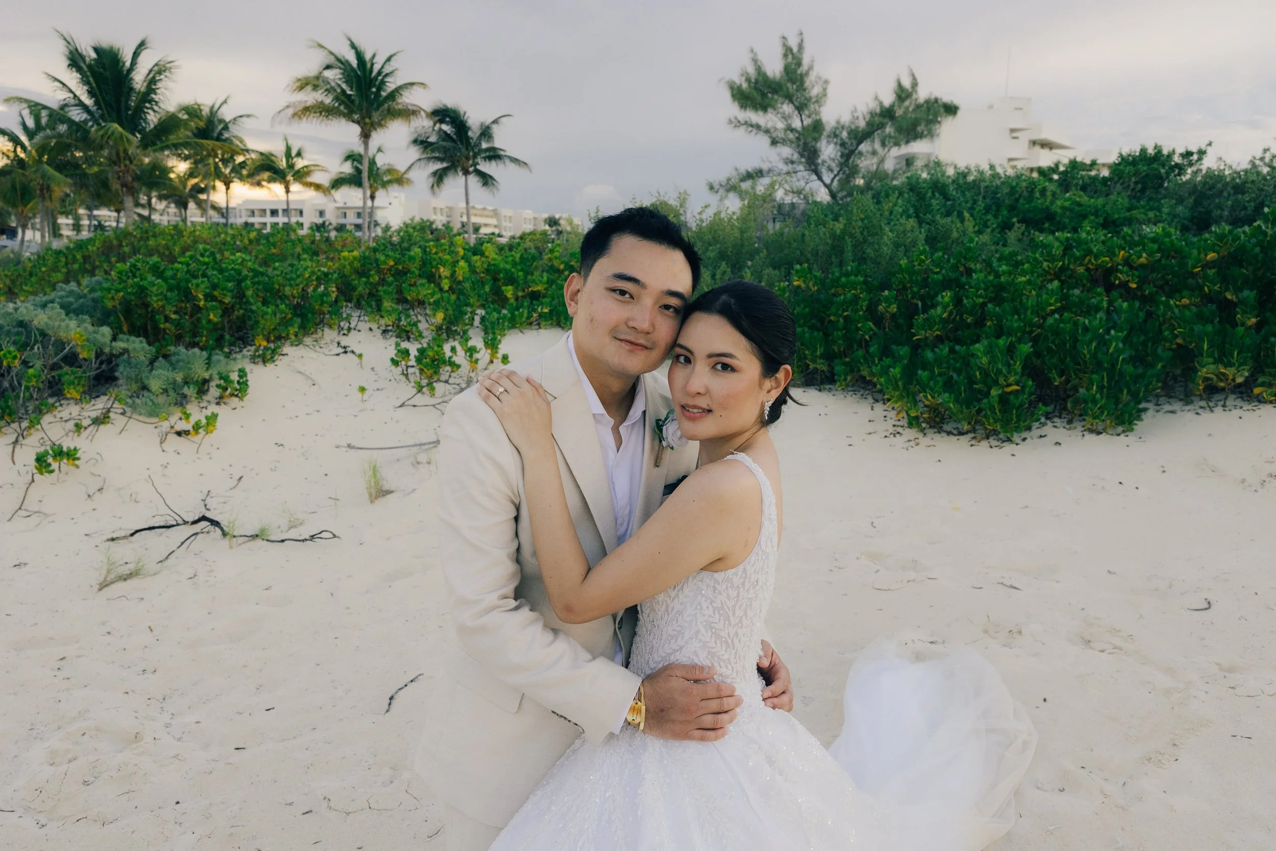 A couple dressed in wedding attire embracing on a sandy beach with greenery and palm trees in the background.