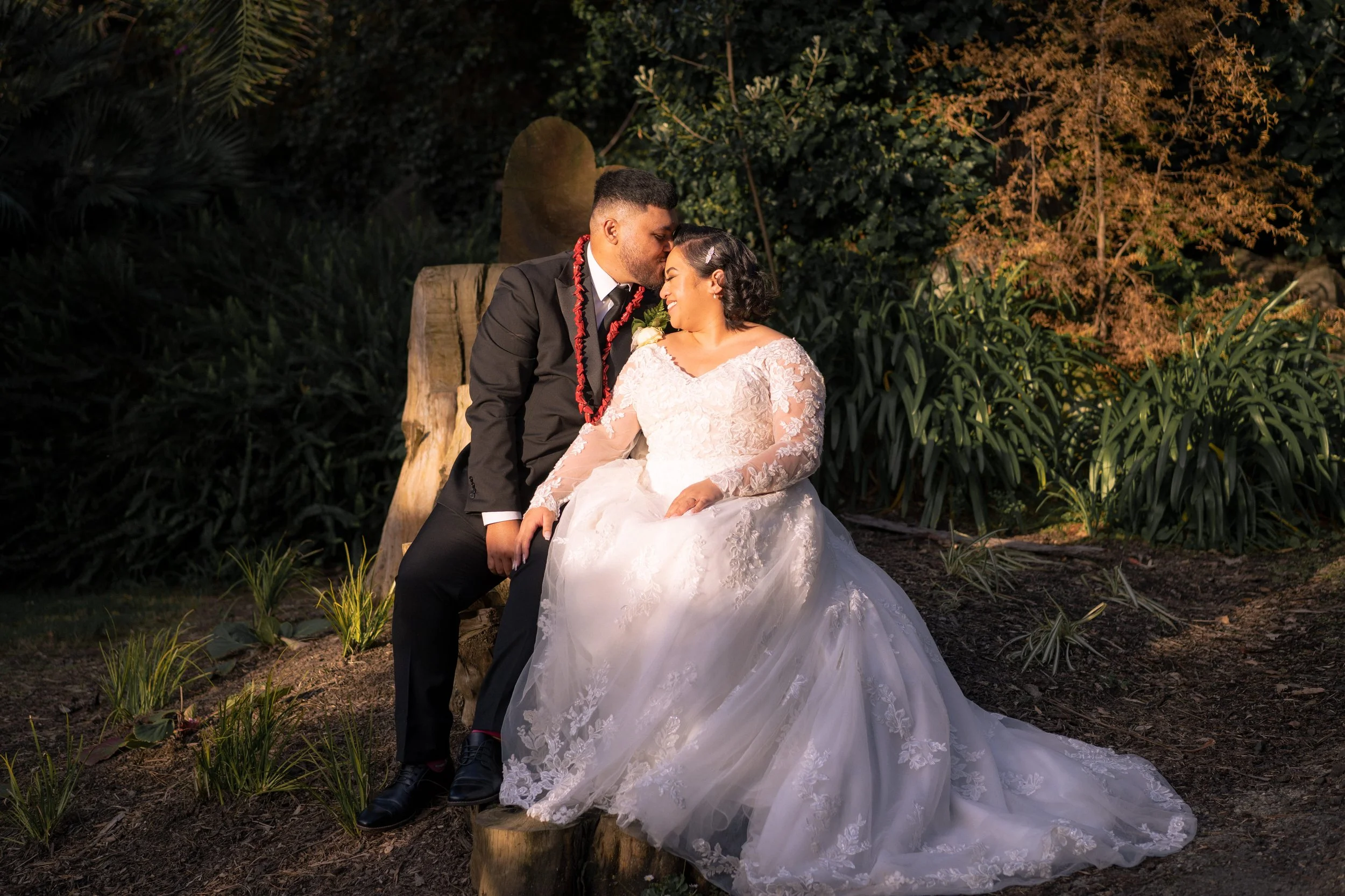 A newlywed couple, dressed in wedding attire, sharing an intimate moment in an outdoor setting at dusk.