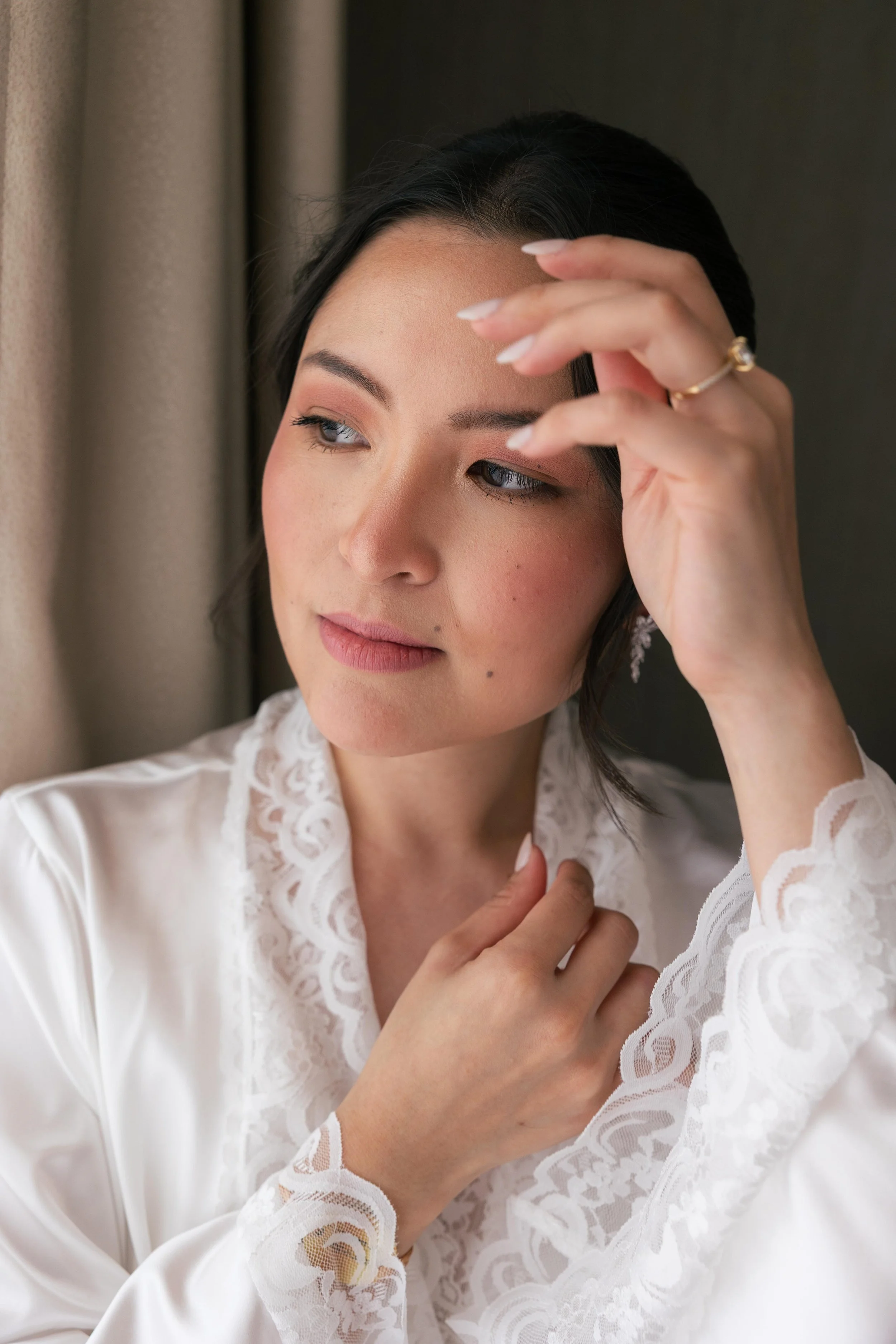 A woman with dark hair and fair skin touching her forehead, wearing a white lace robe, with makeup and a ring, standing near a window with beige curtains.