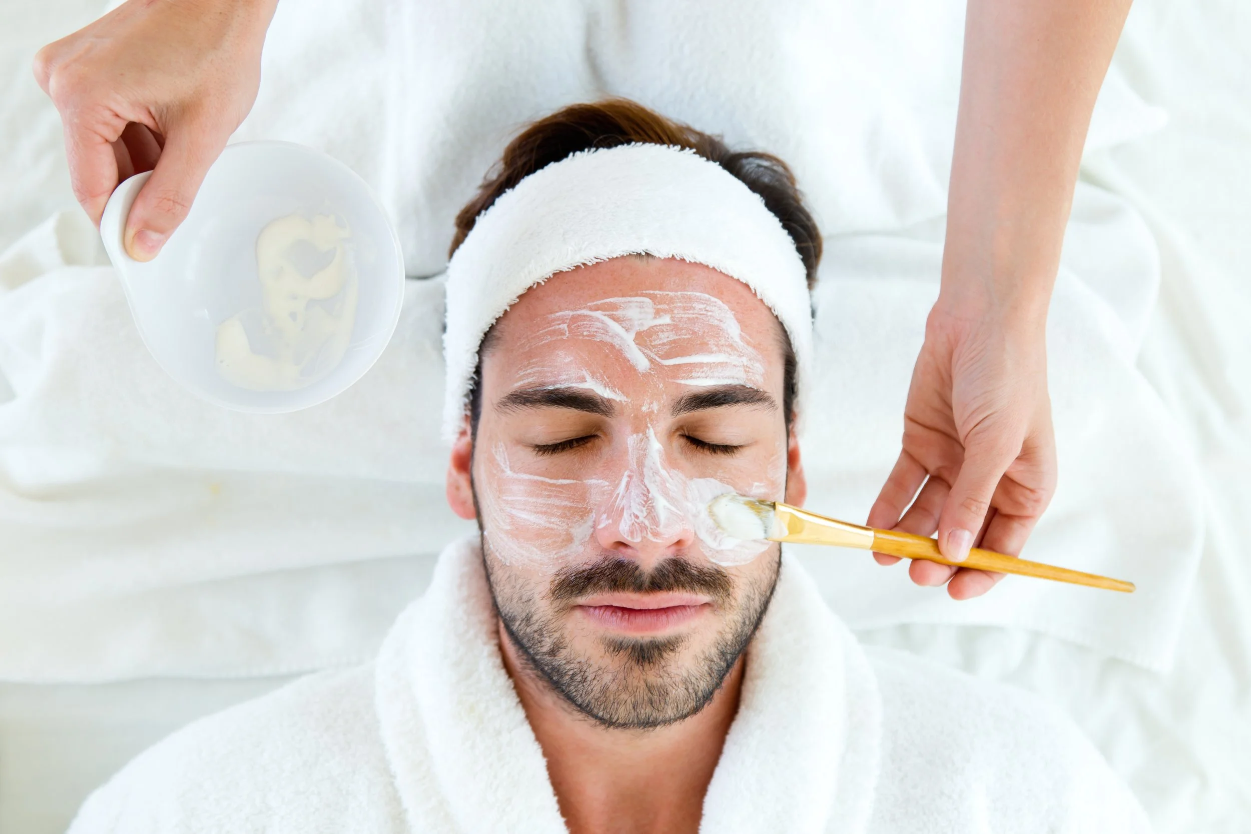 A man with a white headband and towel around his shoulders lying on a spa bed with eyes closed, getting a facial treatment. Aesthetician applying facial mask with a brush, and holding a small dish with cream or mask on it.