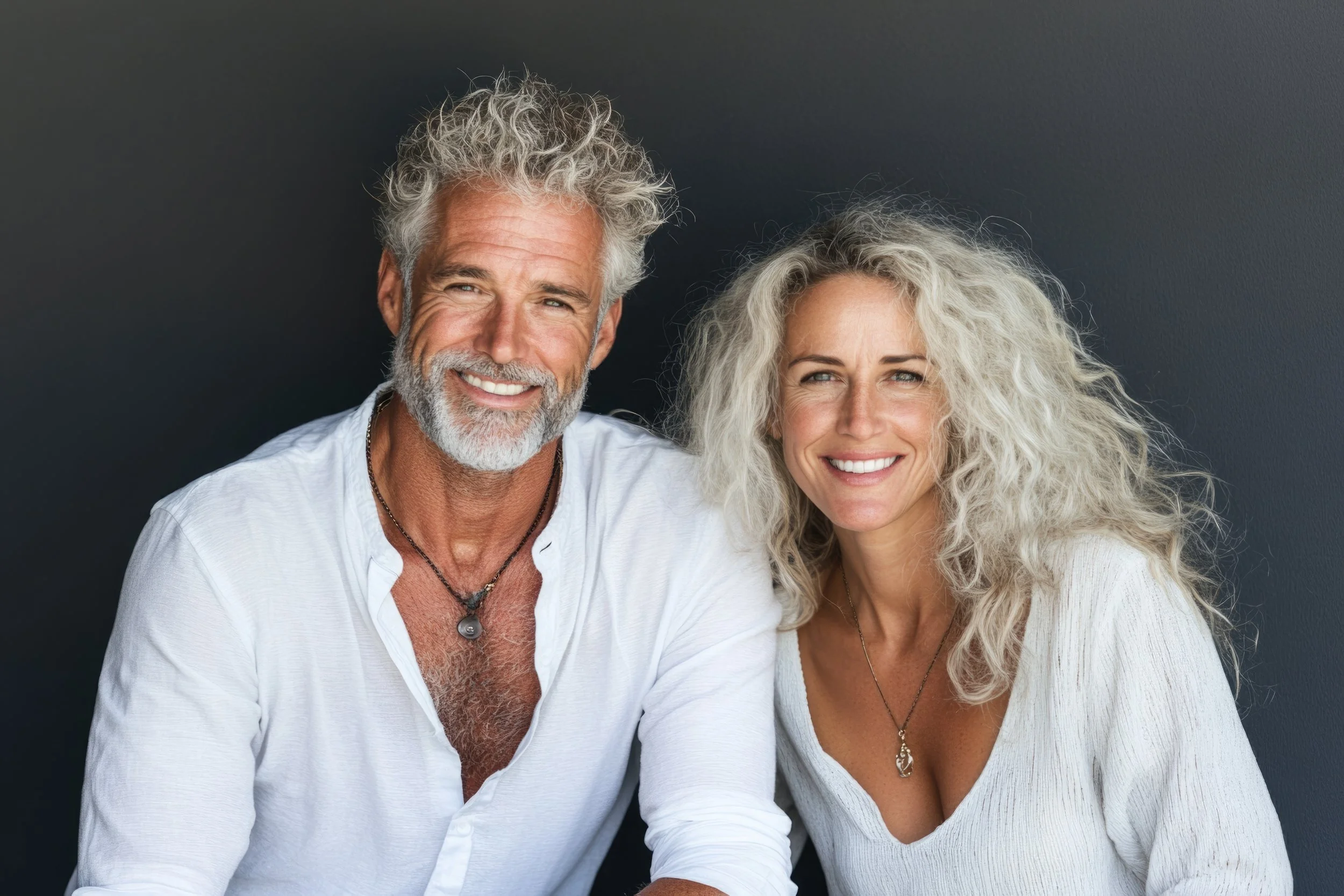 Smiling older man with gray hair and beard wearing a white shirt, sitting next to smiling older woman with curly gray hair wearing a white top, against a dark background.
