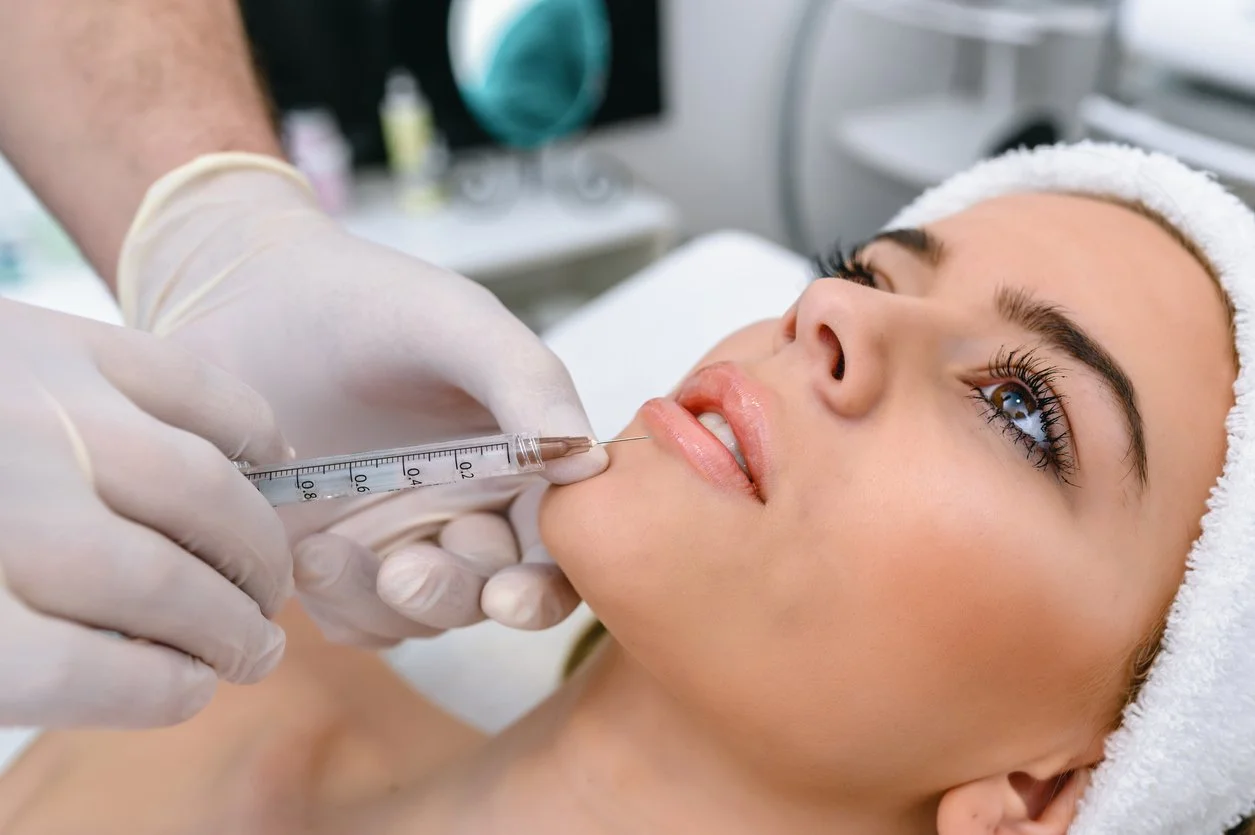 A woman lying down receiving a cosmetic lip augmentation injection from a medical professional in a clinical setting.