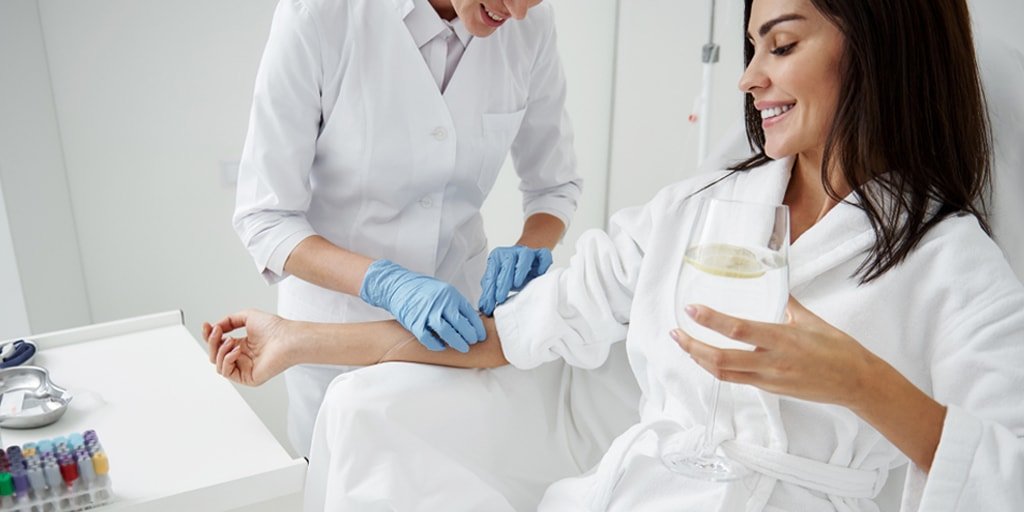 A woman sitting in a hospital bed receiving an intravenous injection from a nurse. The woman is holding a glass of water and smiling.