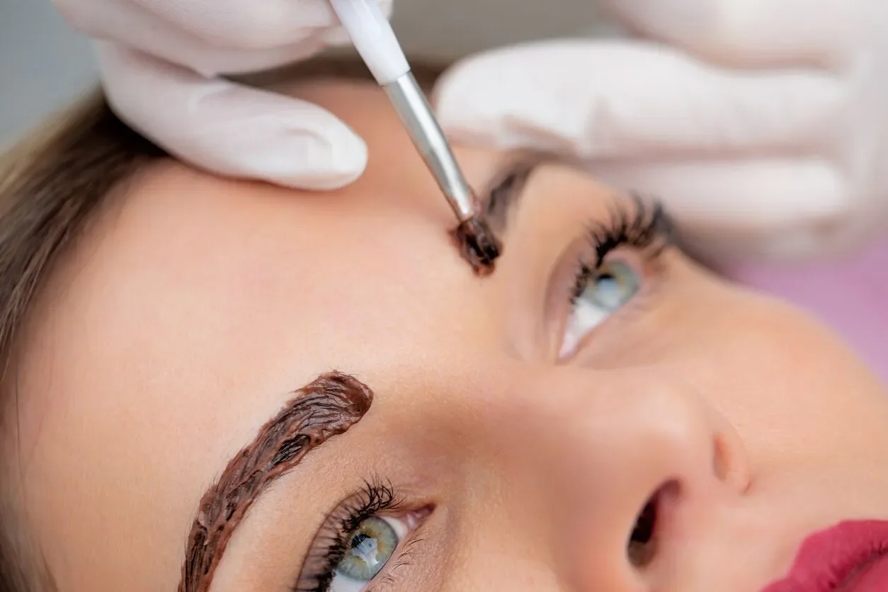 A person lying down with their eyes open and eyebrows being tattooed or shaped by a professional using a small tool, while wearing gloves.