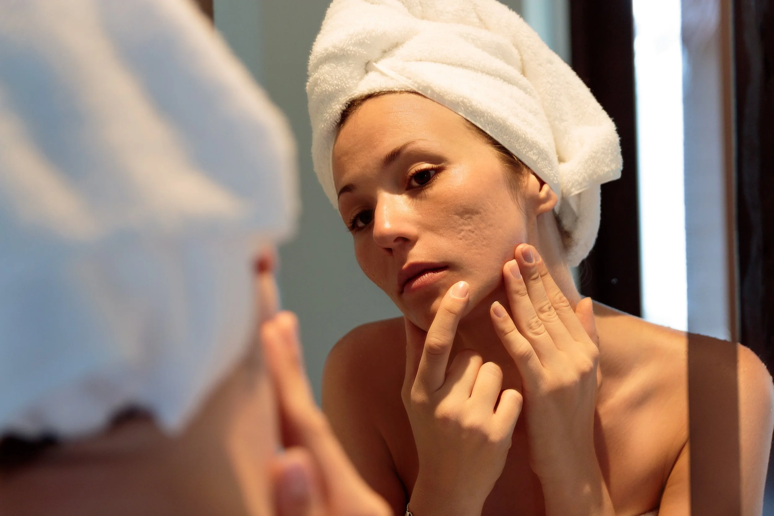 Woman with a towel on her head looking at her reflection in a mirror, touching her face