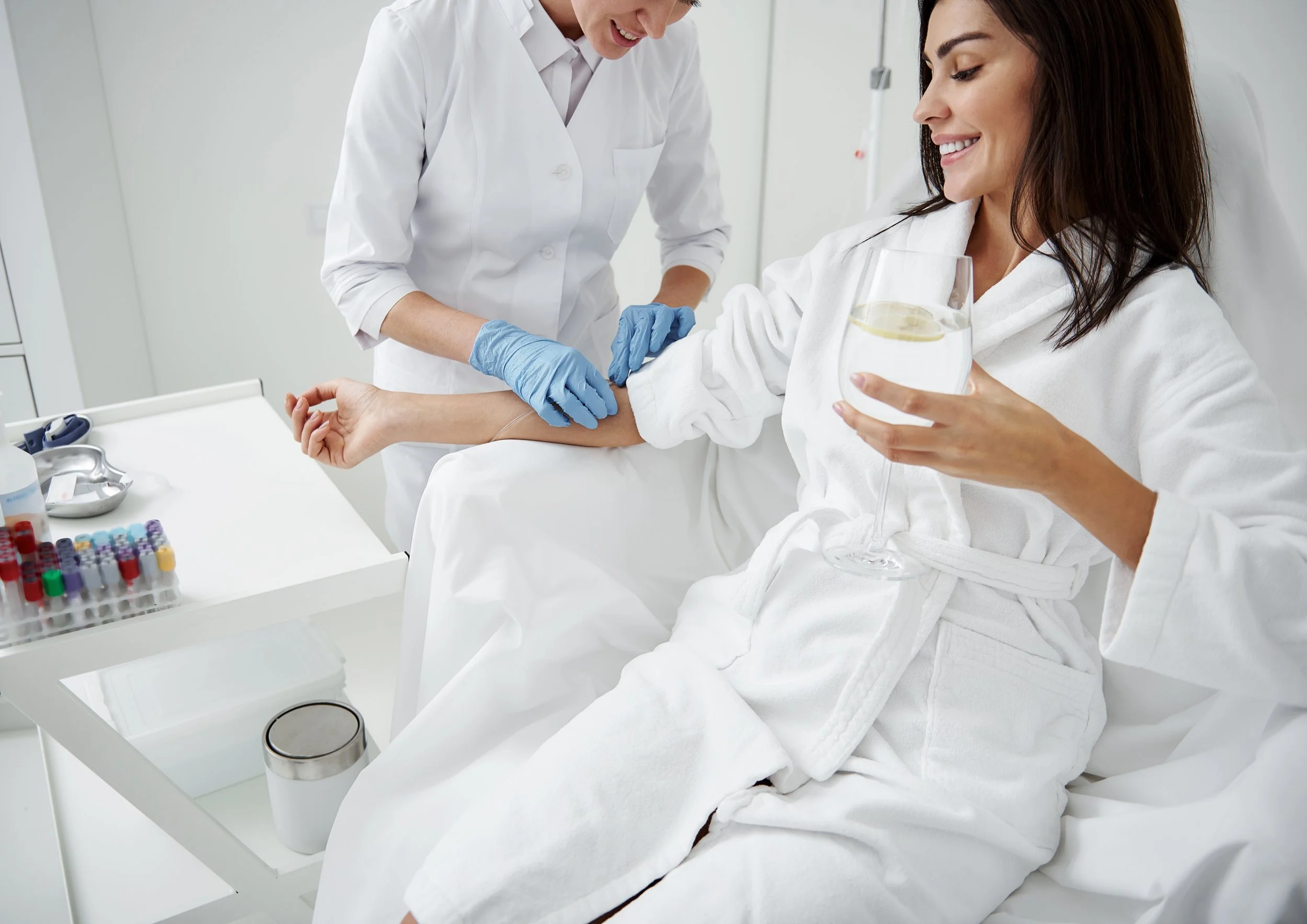 A woman in a white bathrobe sitting on a hospital bed, holding a glass of water with lemon, smiling while a healthcare professional prepares to draw her blood.