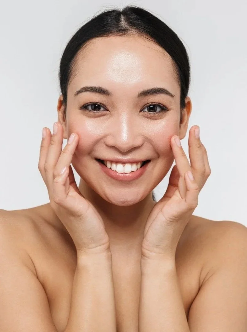 A smiling woman with dark hair pulled back, touching her face with both hands, looking directly at the camera against a plain light gray background.