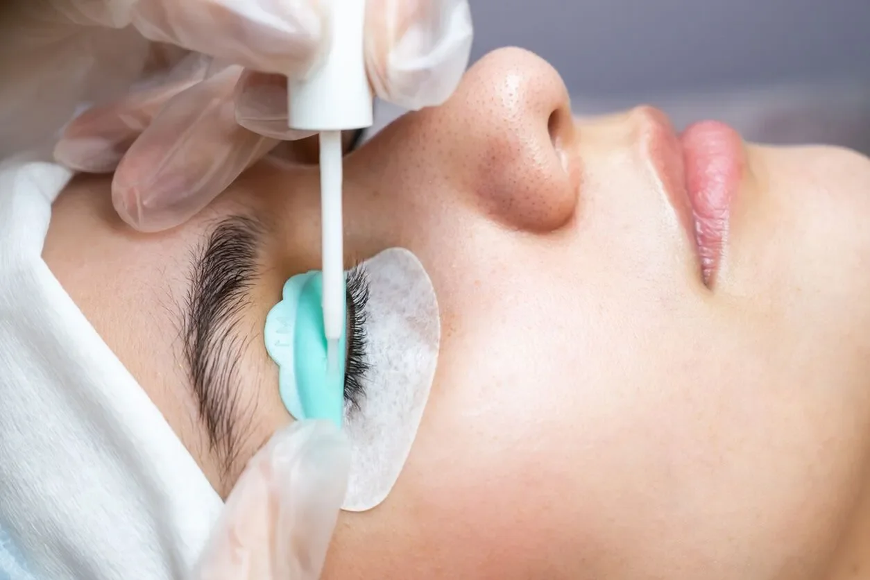 Close-up of a person receiving an eyelash extension treatment, lying down with eye pads under their eyes, while a technician applies lash adhesive with a tool.