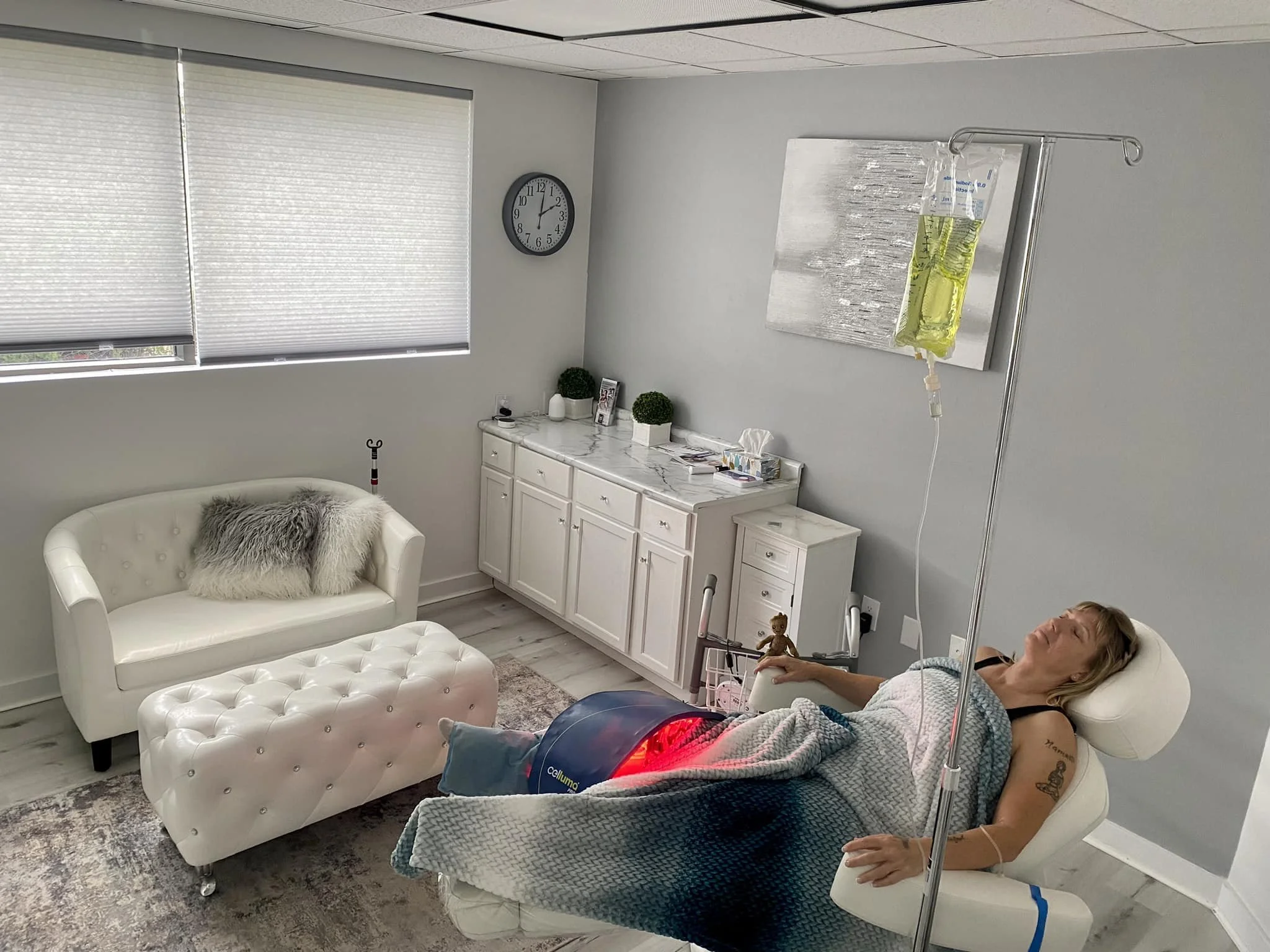 A woman lying on a medical chair receiving IV therapy in a modern, clean clinic room. She appears relaxed with her eyes closed, and an IV bag hangs on a stand next to her. The room has a white couch, a marble-topped cabinet, and a clock on the wall.