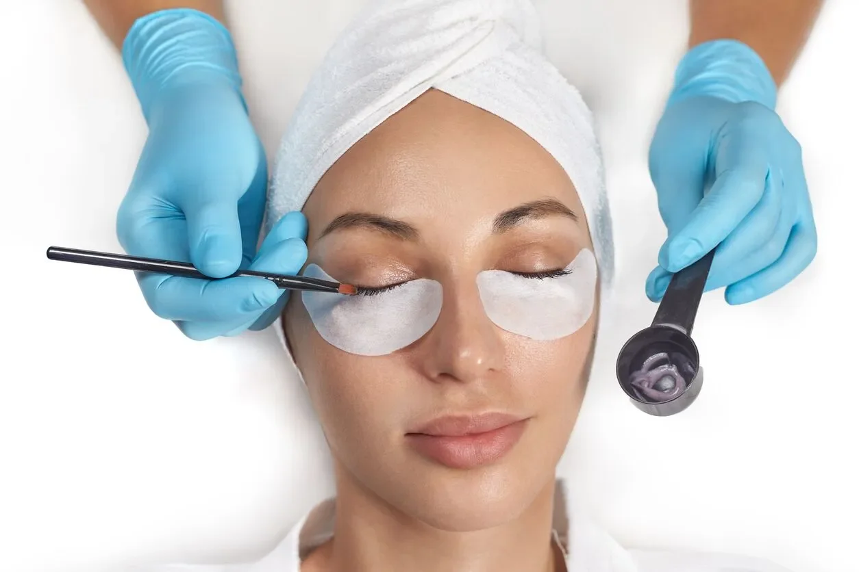 A woman with eyes closed receiving a facial treatment with eye masks, while a professional applies makeup or skincare with a brush and a spatula, both wearing blue gloves.