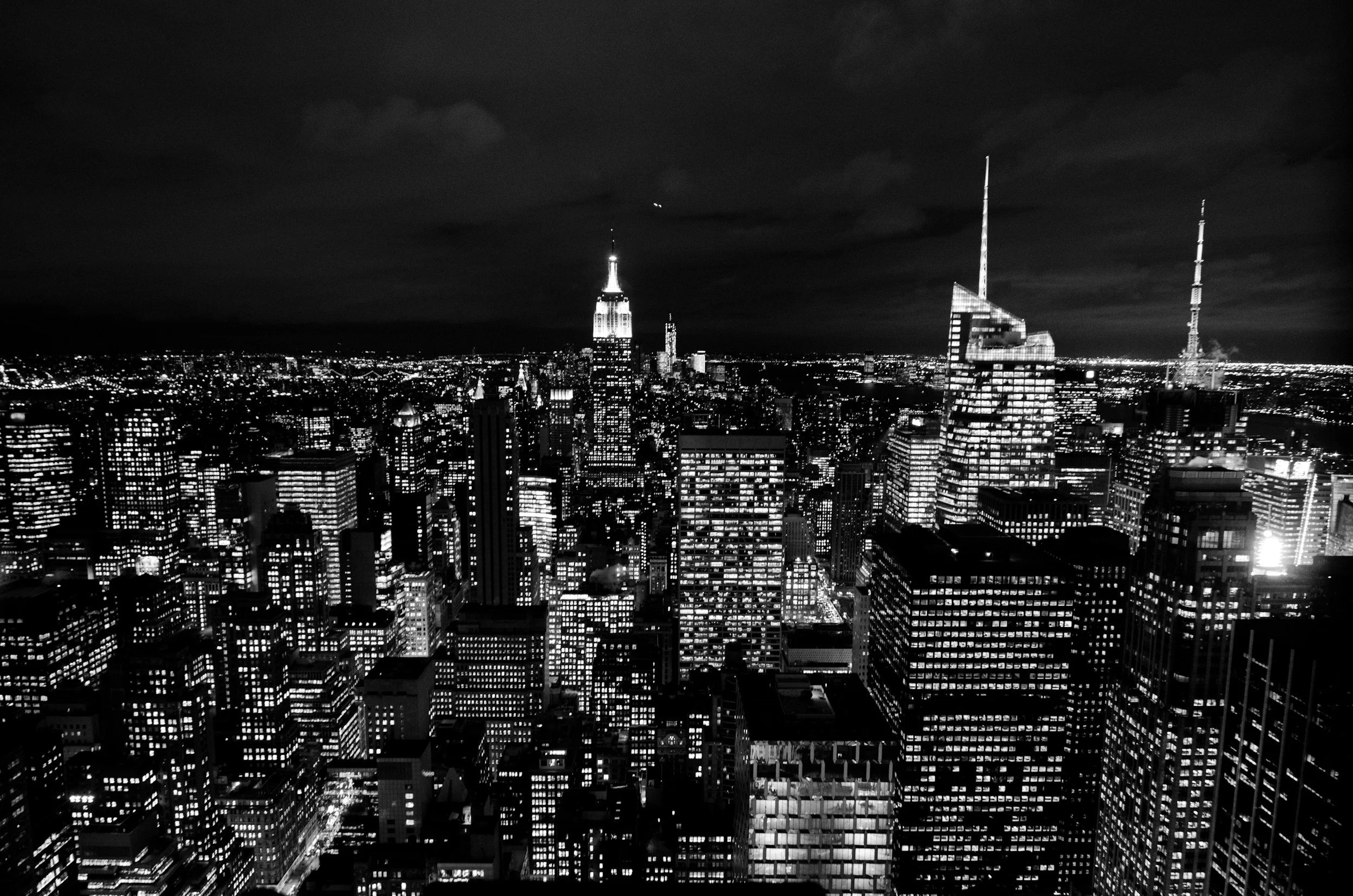 Nighttime view of the New York City skyline with illuminated skyscrapers including the Empire State Building and One Vanderbilt building, in black and white.