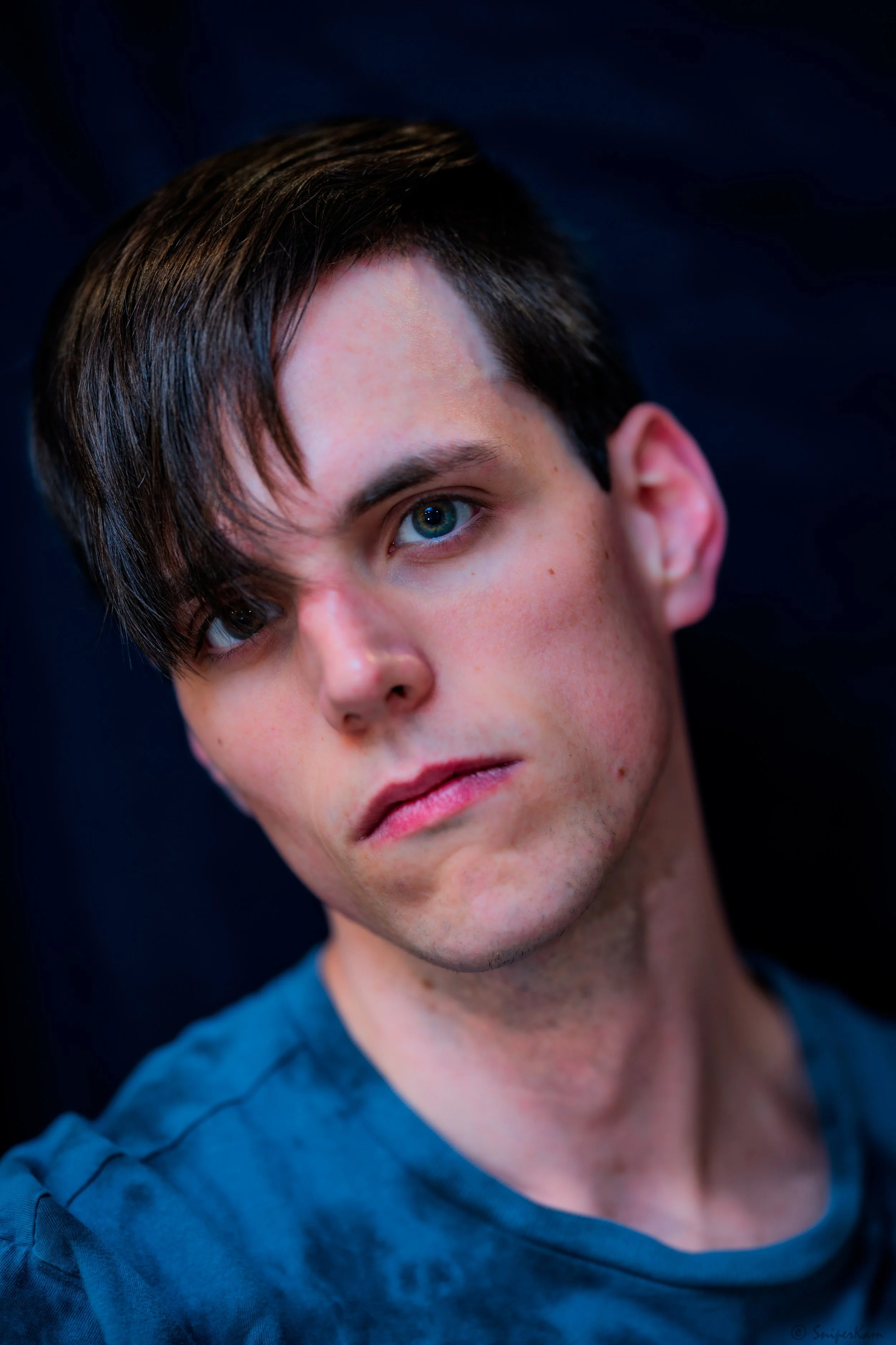 Close-up of a young man with dark hair and blue eyes, wearing a blue shirt, looking to the side against a dark background.