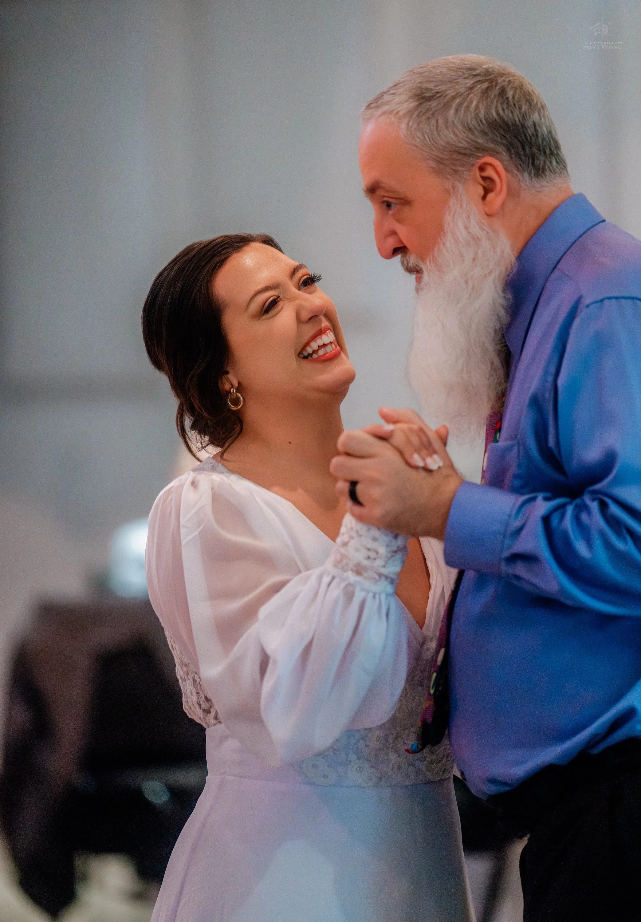 A woman with dark hair smiling and holding hands with a man who has a gray beard and mustache, and gray hair. They are dancing closely in a joyful moment. The woman is wearing a white dress with lace details, and the man is wearing a blue shirt. The 