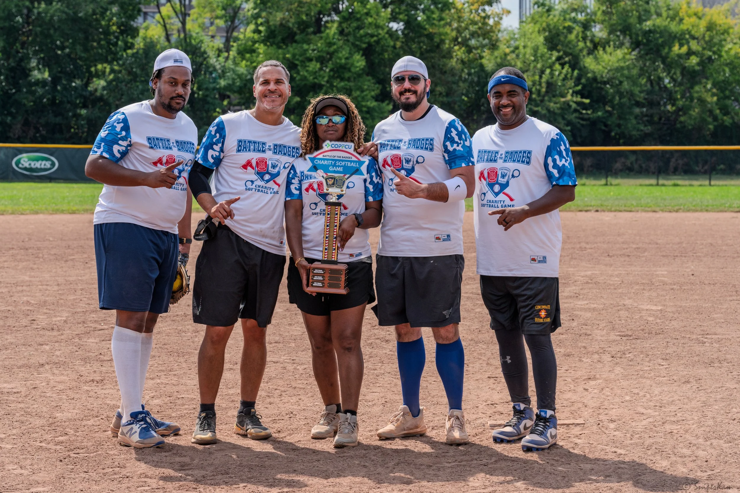 Group of five people holding a softball trophy, standing on a softball field, wearing matching white and blue shirts with a tournament logo, smiling and pointing at the trophy.