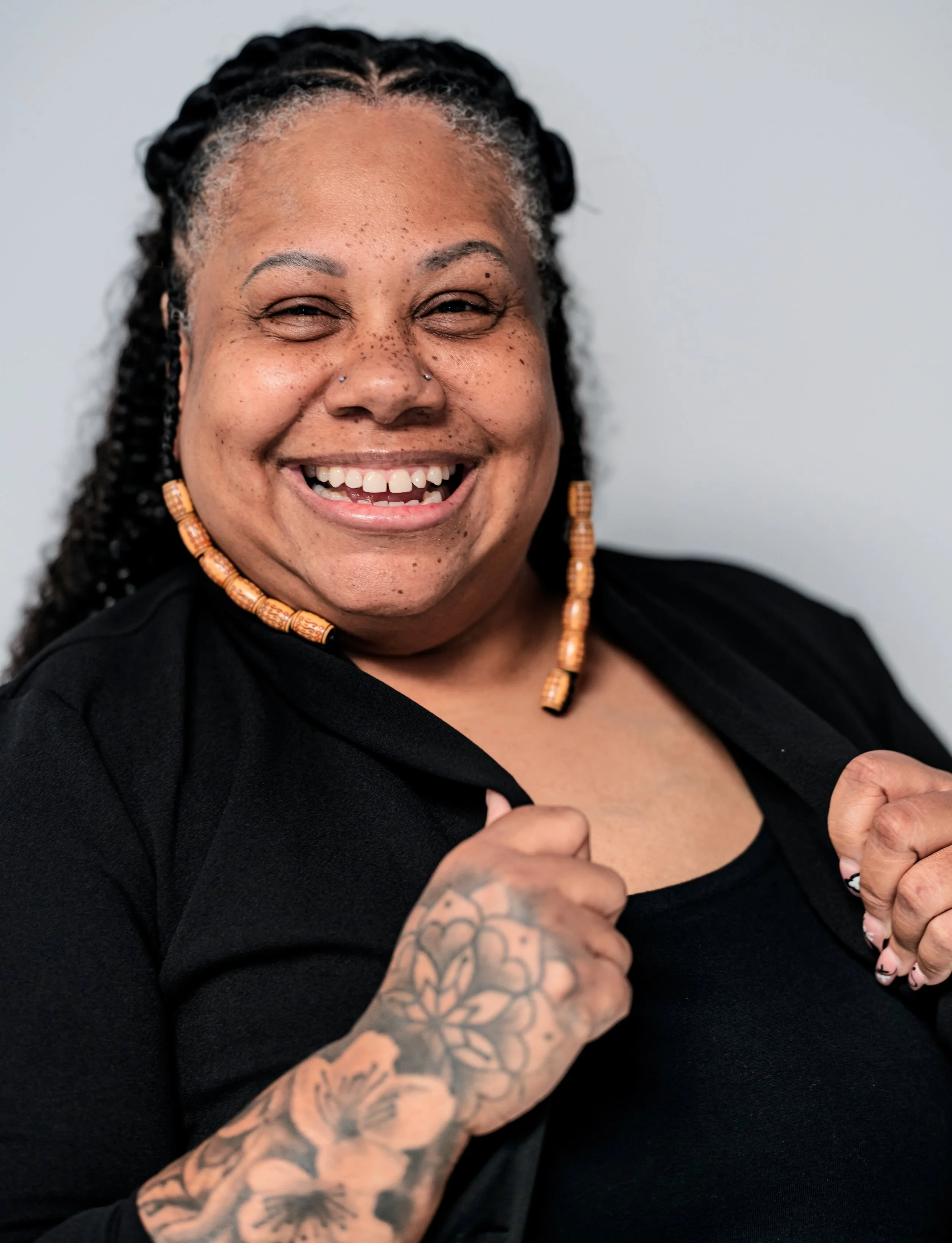 Close-up of a smiling woman with black hair styled in braids, wearing orange beaded earrings, a black top, and a tattoo on her left forearm, holding open her black jacket.
