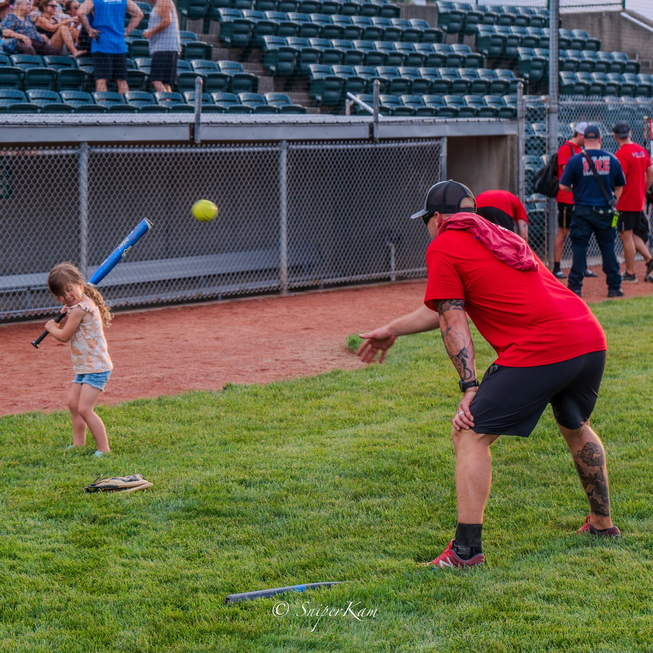 A young girl swinging a baseball bat, preparing to hit a pitched ball, with a man in a red shirt and black shorts ready to catch or throw the ball back. They are on a grassy field near a baseball diamond, with empty bleachers and a chain-link fence i