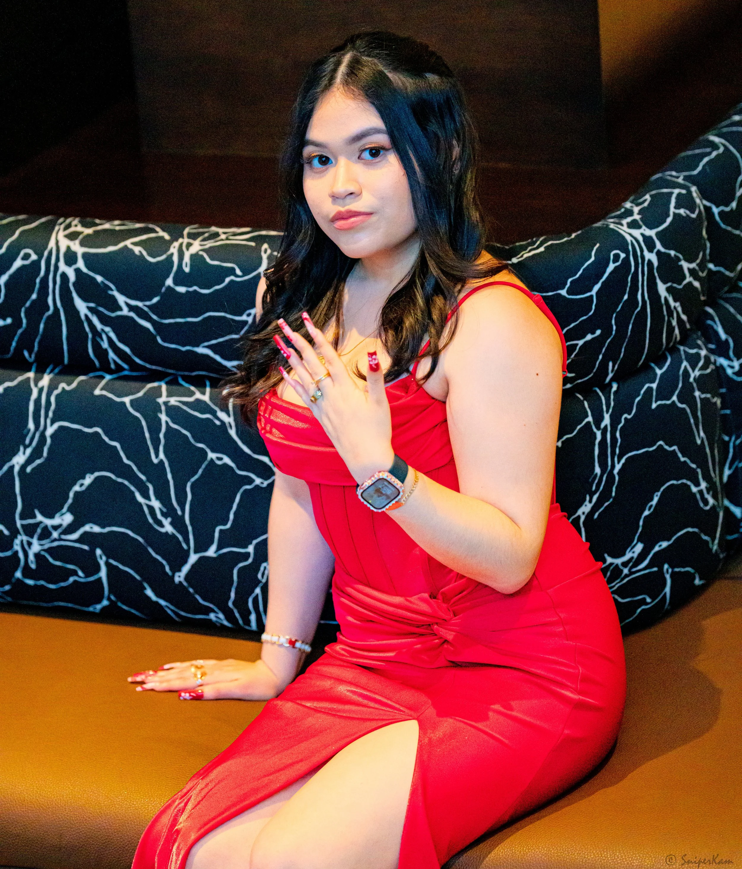 A woman in a red dress sitting on a brown bench with a black and white patterned backrest behind her, with dark hair, makeup, and jewelry.