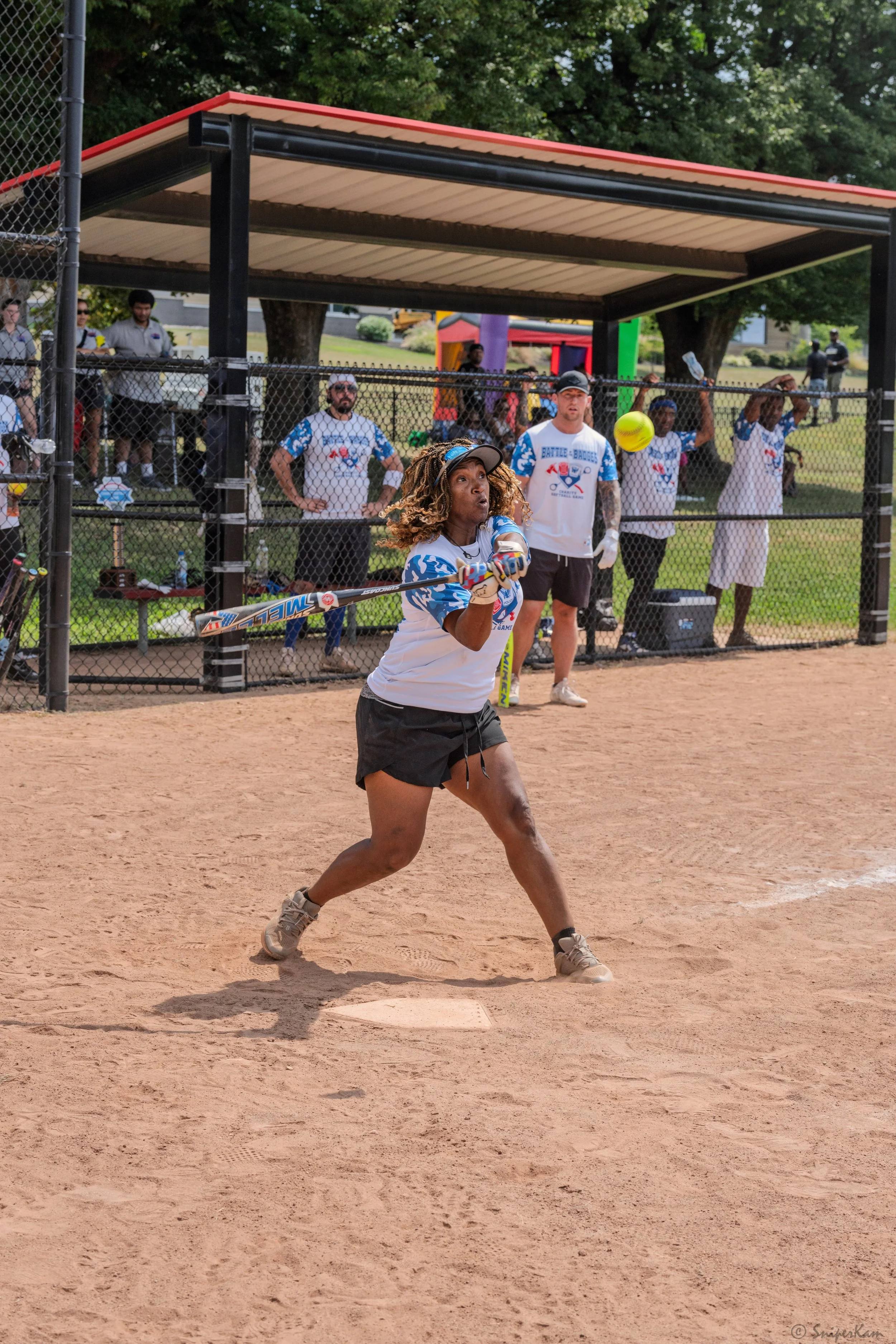 Woman batting in a softball game on a dirt field, with a fence and spectators in the background.