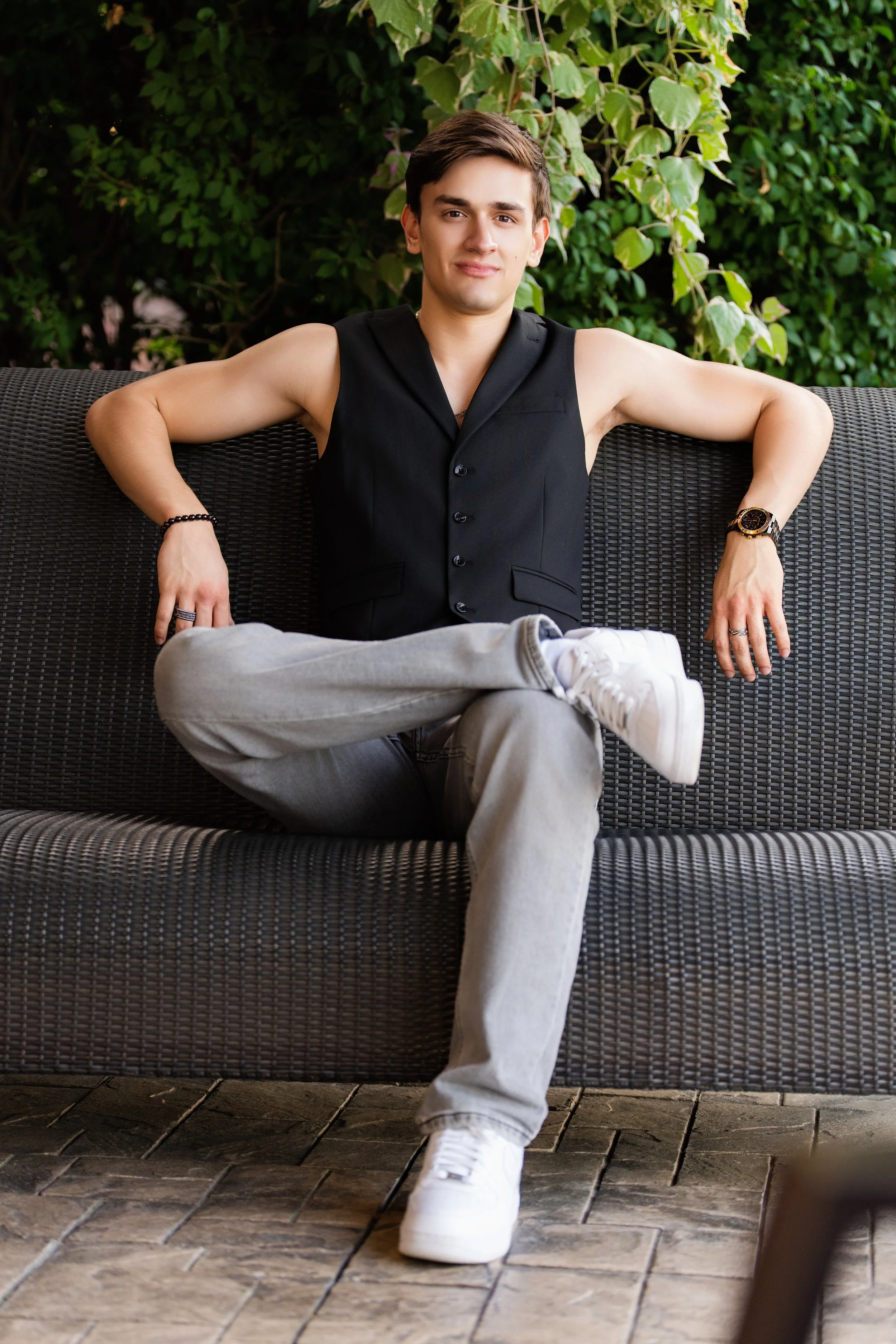 A young man with short brown hair, sitting on a dark outdoor sofa with a background of green leafy plants. He is wearing a sleeveless black vest, gray jeans, white sneakers, a bracelet, and a watch. He has a relaxed pose with one leg crossed over the
