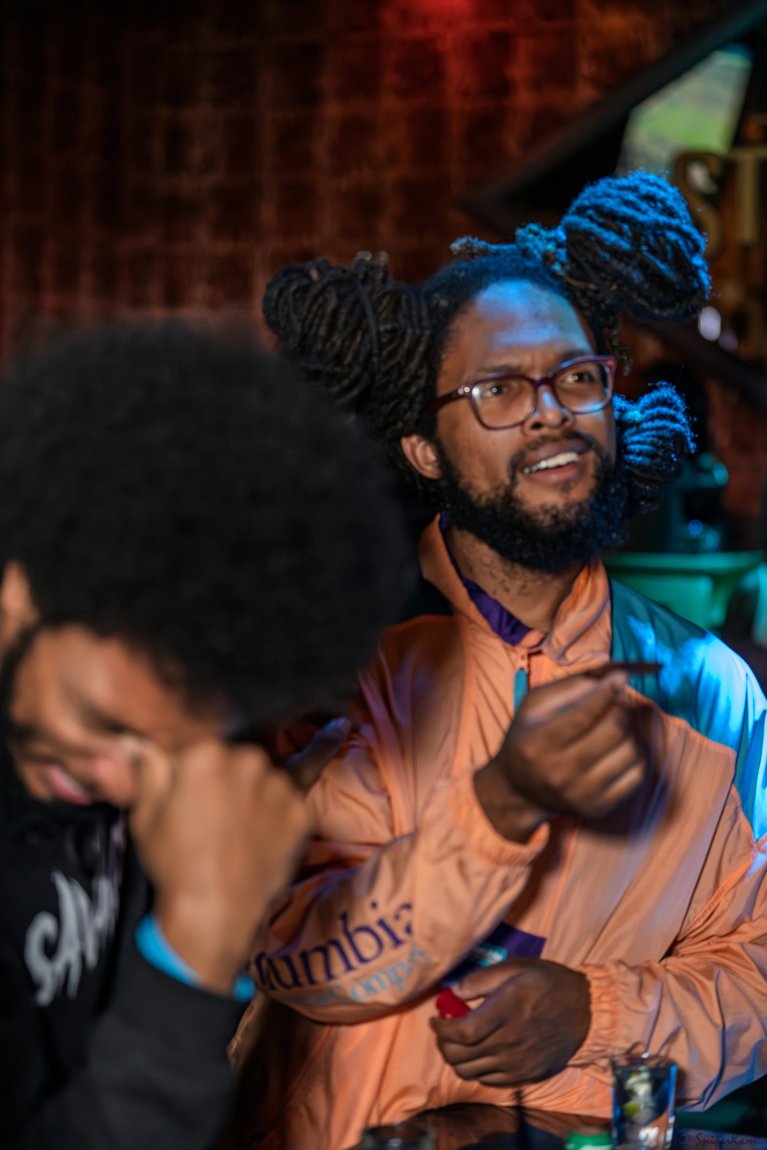 Two men engaged in conversation, one with glasses and dreadlocks styled into large buns, wearing a colorful jacket, and gesturing with his hand, while the other, with glasses and facial hair, appears to be listening.