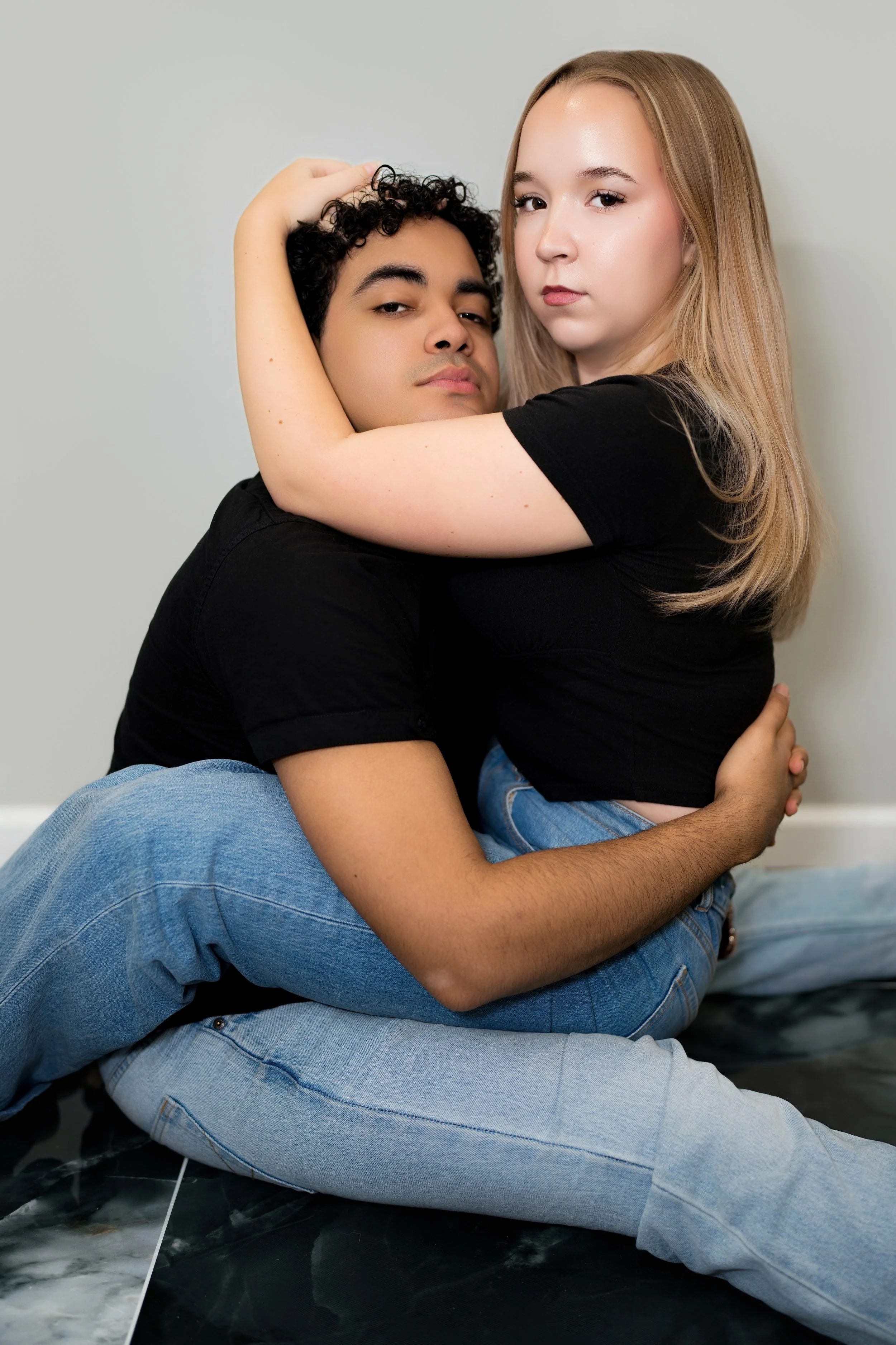 A young woman and man sitting closely with the woman embracing the man around his neck, both looking serious. They are wearing black shirts and blue jeans, seated on a dark marble floor against a plain, light wall.