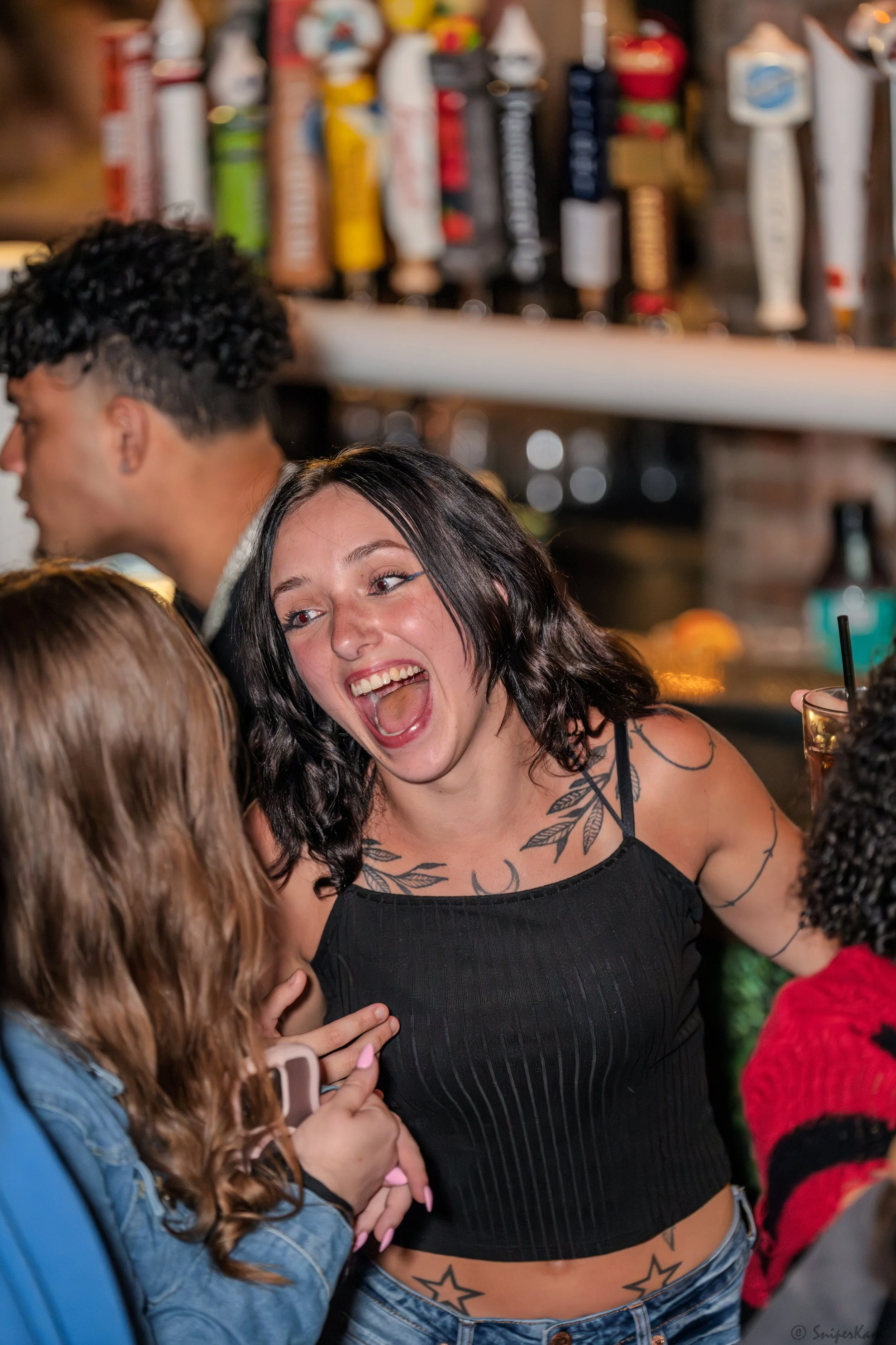 A woman with black hair, tattoos on her shoulders and stomach, wearing a black sleeveless top, is smiling and interacting with friends at a bar or social gathering.