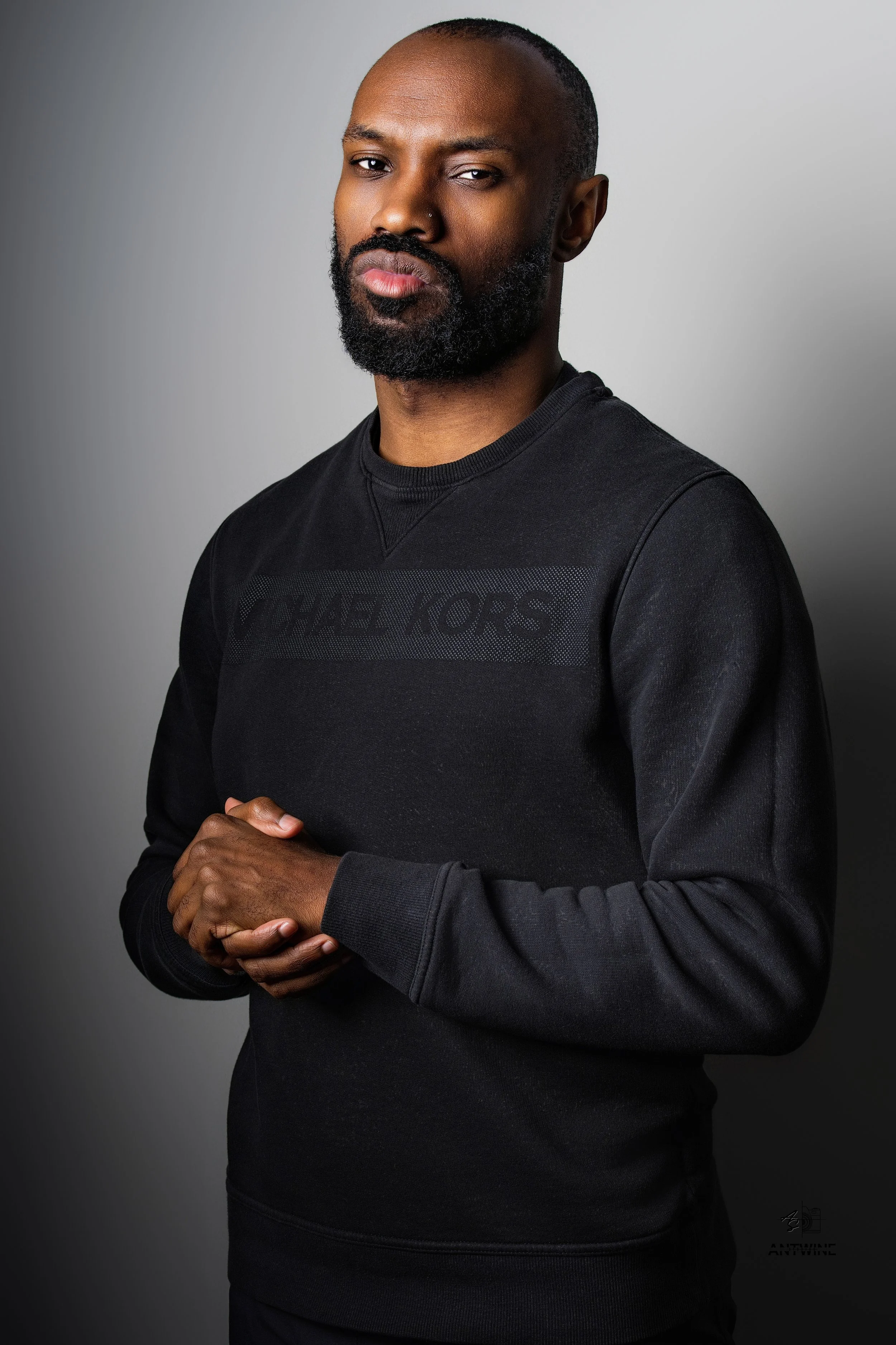A man with a beard and short hair wearing a black Michael Kors sweatshirt, standing against a gray background, looking at the camera with a serious expression and clasping his hands together.