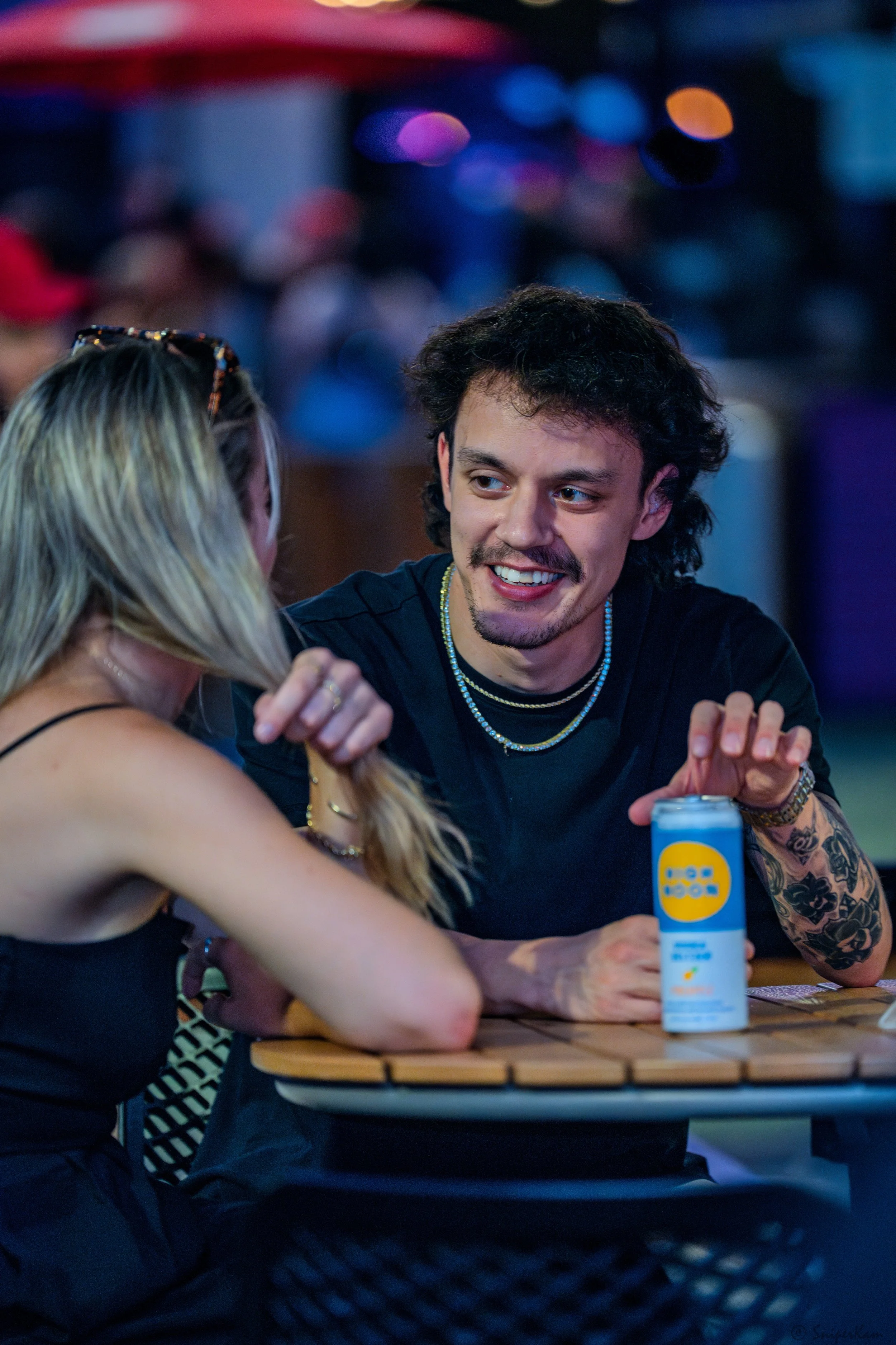 A man and woman sitting at a table, smiling and talking, with a can of beverage on the table, in a lively outdoor setting with blurred colorful lights in the background.