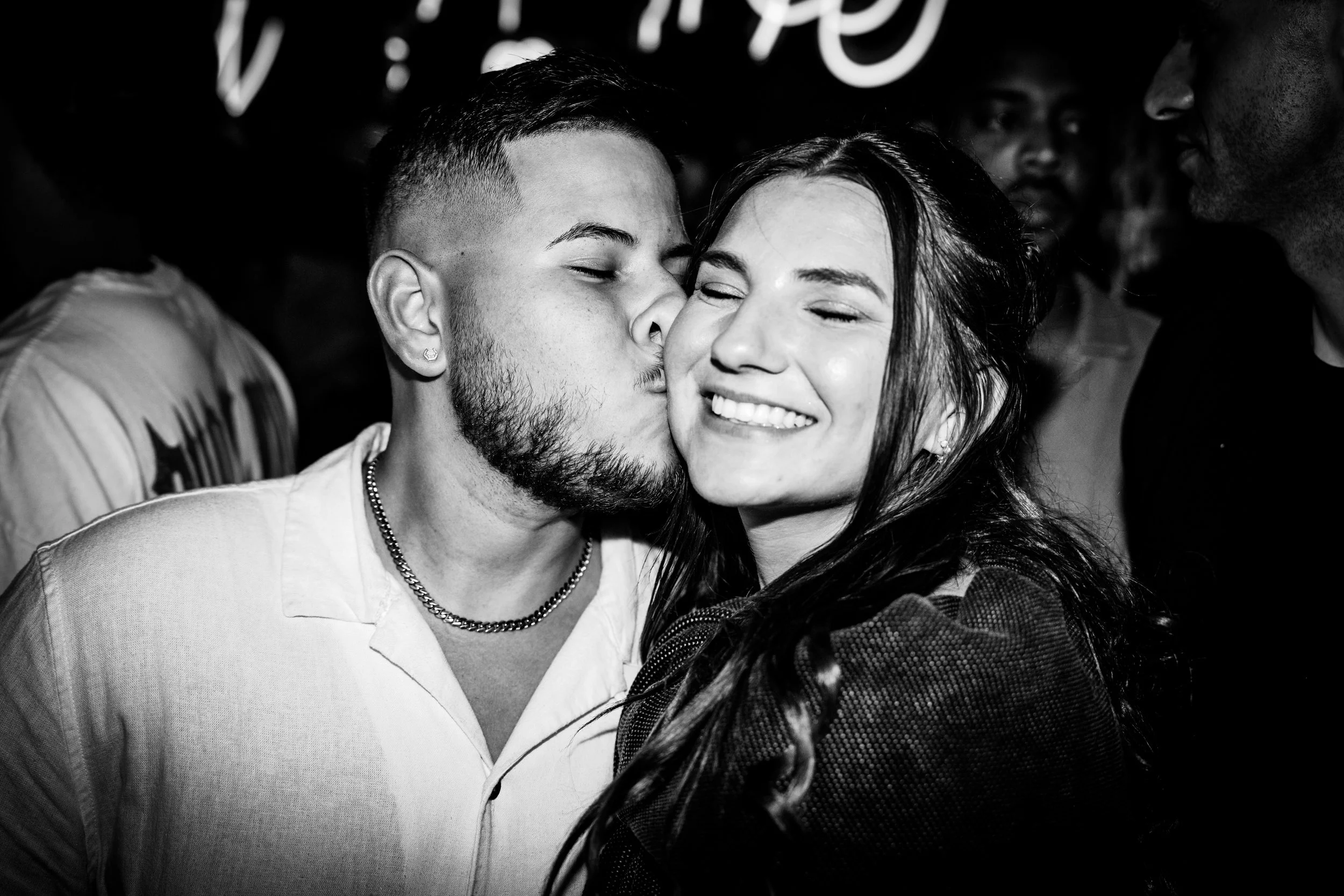 A man kissing a woman on the cheek at a social event, smiles with closed eyes, surrounded by other people, in black and white.